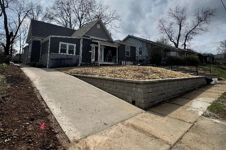 A residential house with a sloped sidewalk in the foreground, a retaining wall, bare trees, and an overcast sky.