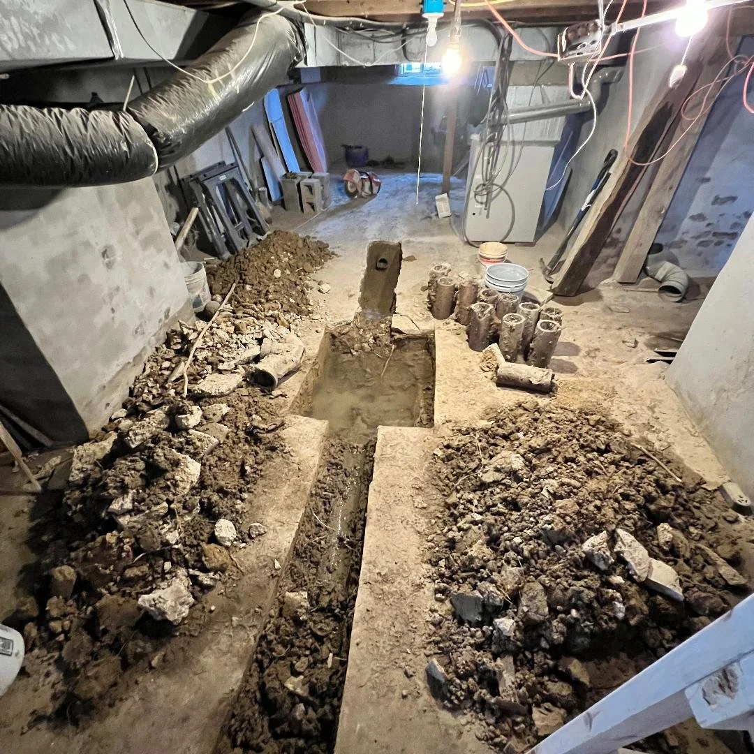 Construction site with a large rectangular hole in the ground, surrounded by piles of dirt and bricks, and construction tools and materials scattered around.