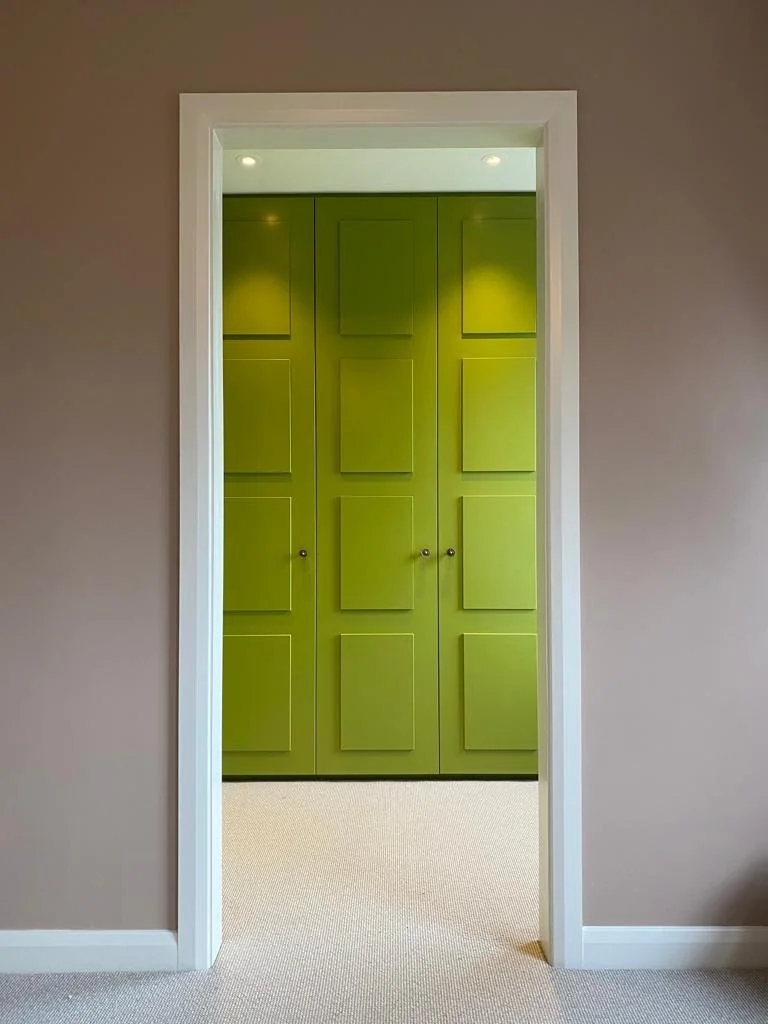 A glimpse through to a dressing room where the cupboard doors are painted a striking acid green. The cupboard doors have reverse panelling on them with 4 vertical panels on each door and they are finished with brass door handles.