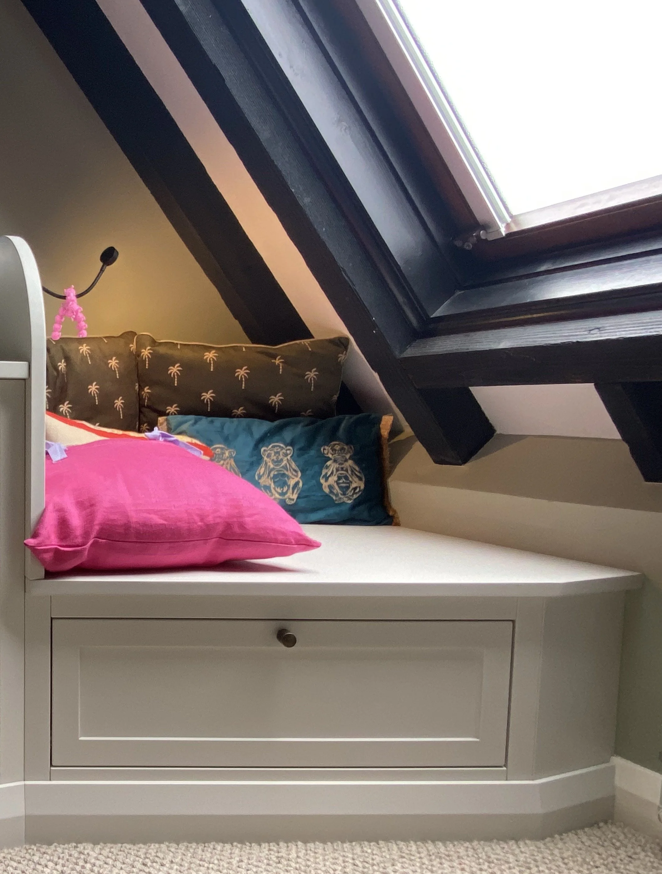 A cosy reading nook seat that cleverly makes the most of an awkward corner between the desk and velux window. There's a shaker style storage drawer beneath it and it's painted in a calming grey with colourful scatter cushions bringing interest and li