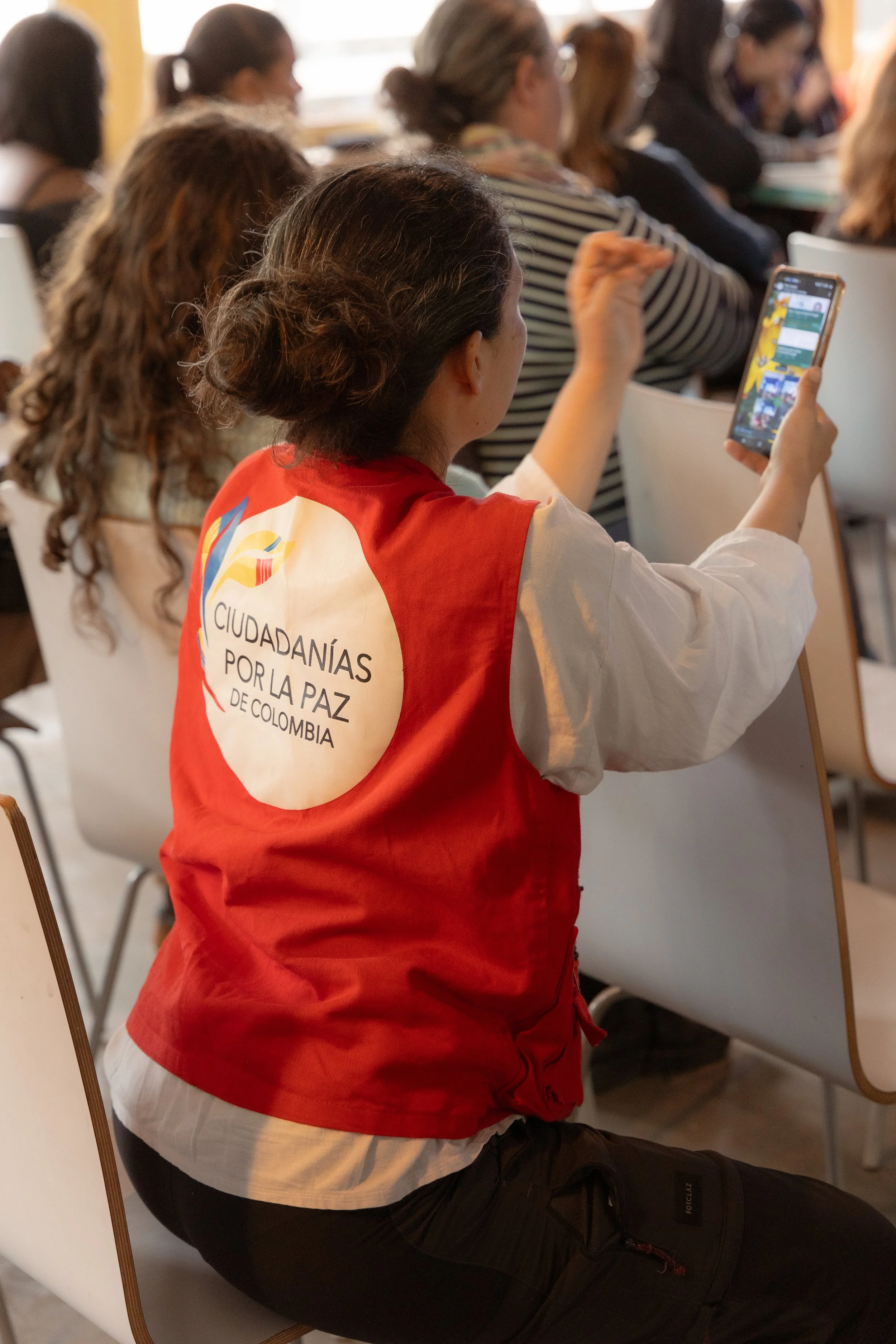 Une femme portant un gilet rouge avec l'inscription « Ciudadanías por la paz de Colombia » assise parmi un groupe de personnes, tenant un téléphone portable et regardant l'écran.
