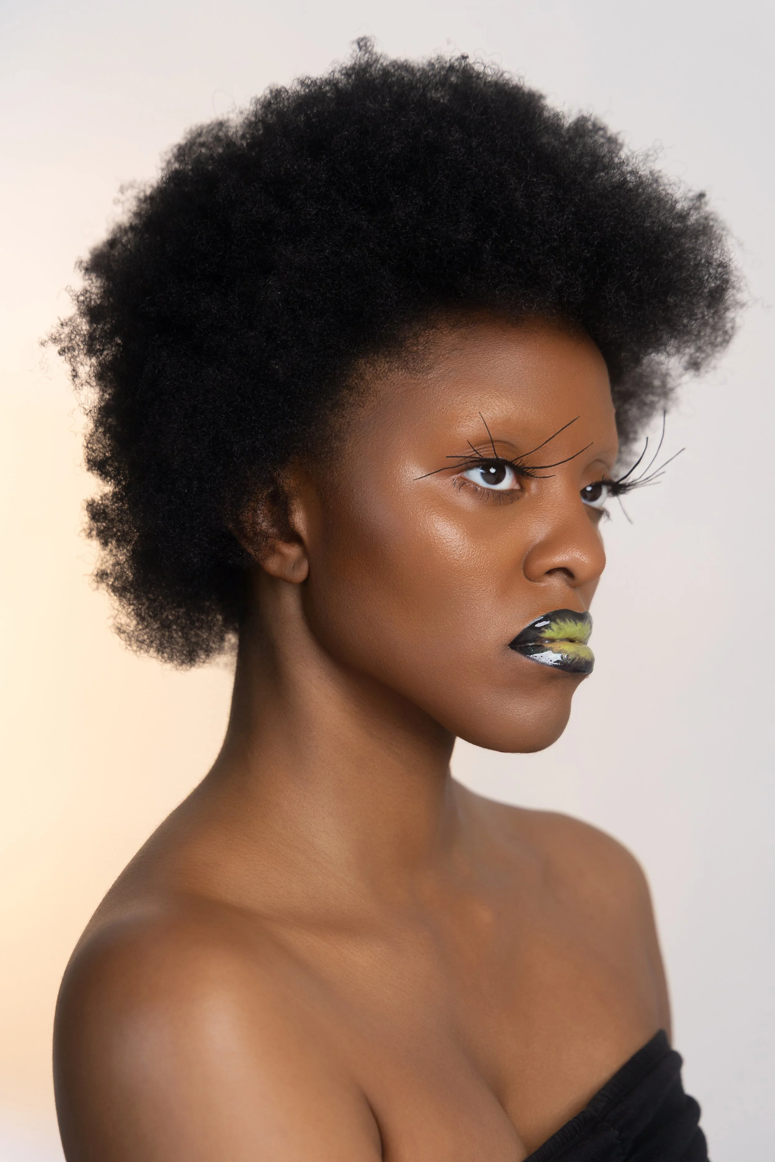 Shooting beauté. Portrait d'une femme noire avec coiffure afro, maquillage artistique, yeux accentués avec des faux cils, lèvres peintes en noir et vert, peau lumineuse, vêtue d'un haut noir hors épaule.