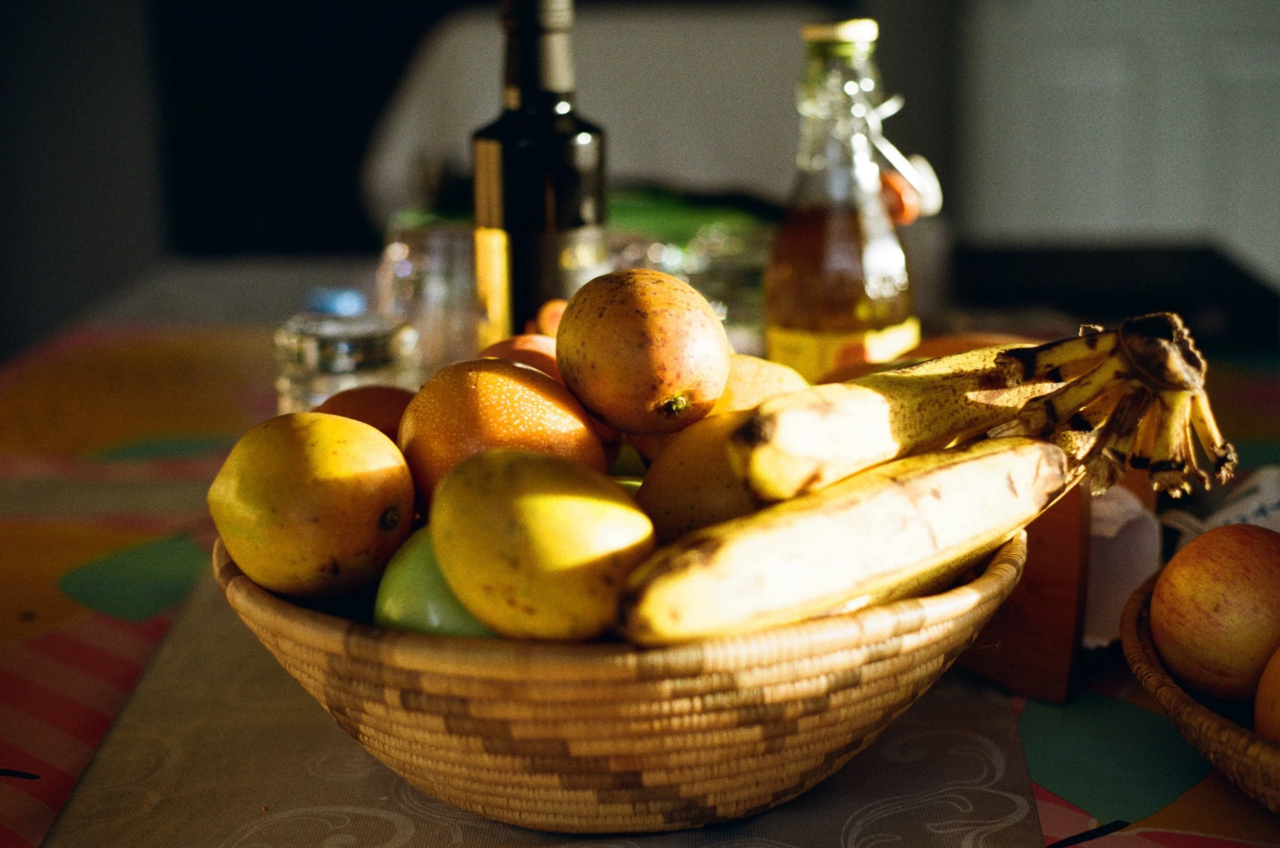 Photo en Colombie. Panier en osier rempli de pommes, bananes, et autres fruits, sur une table avec bouteilles et verres en arrière-plan
