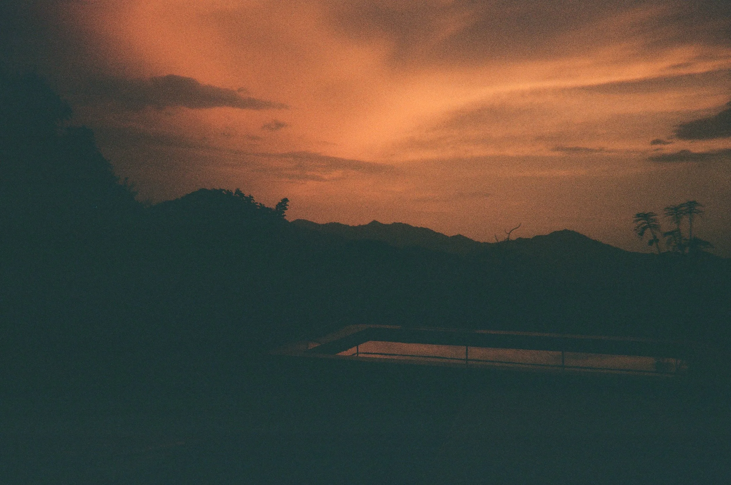 Coucher de soleil avec des nuages orange dans le ciel, silhouettes de montagnes et quelques arbres, vue d'une piscine en contrebas.