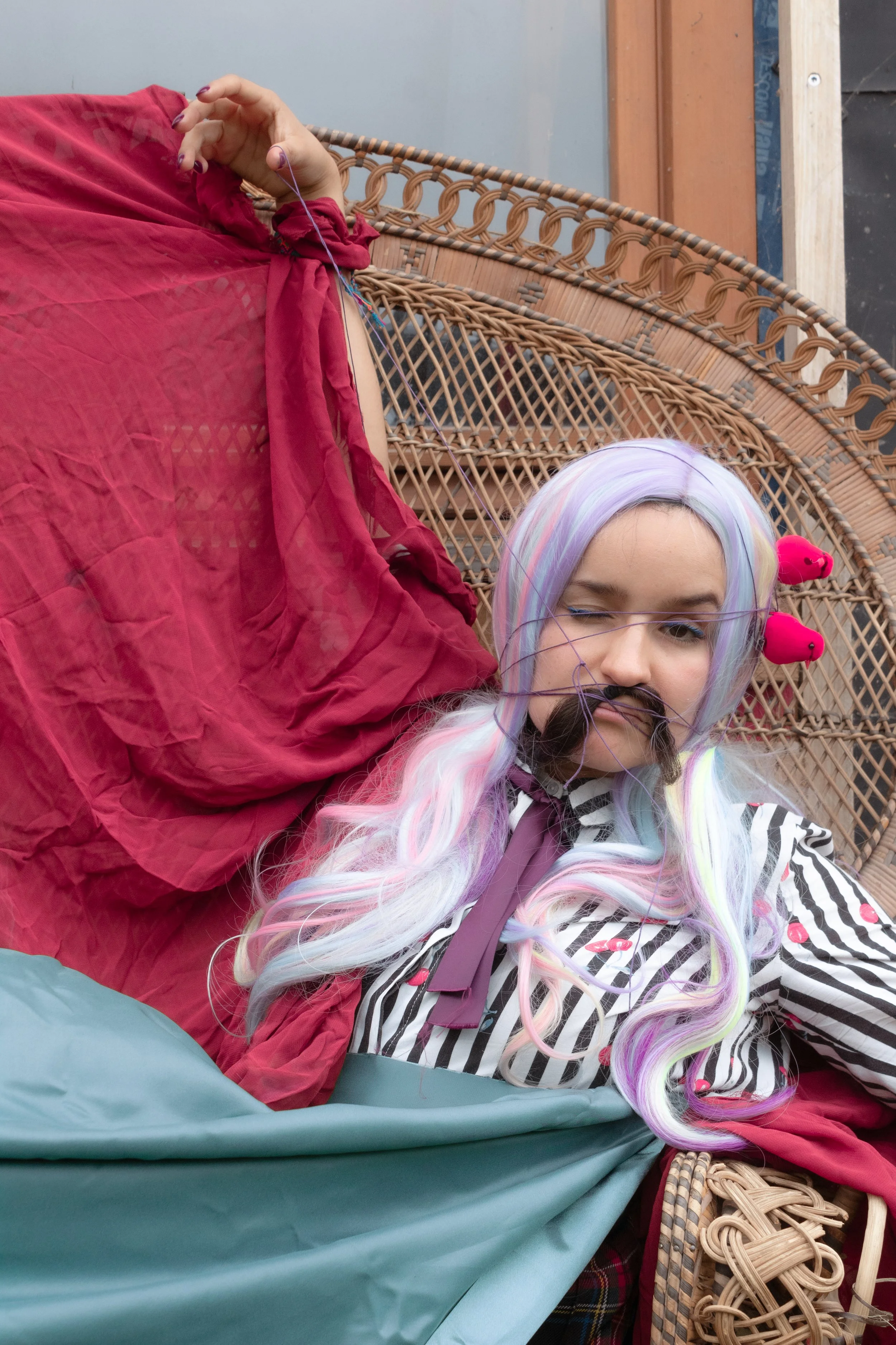 Personne déguisée avec perruque pastel, moustaches en fausse fourrure, coiffée de deux épingles roses, allongée sur un fauteuil en rotin, portant une chemise rayée avec des cerises en broderie et un nœud violet, dans une ambiance colorée