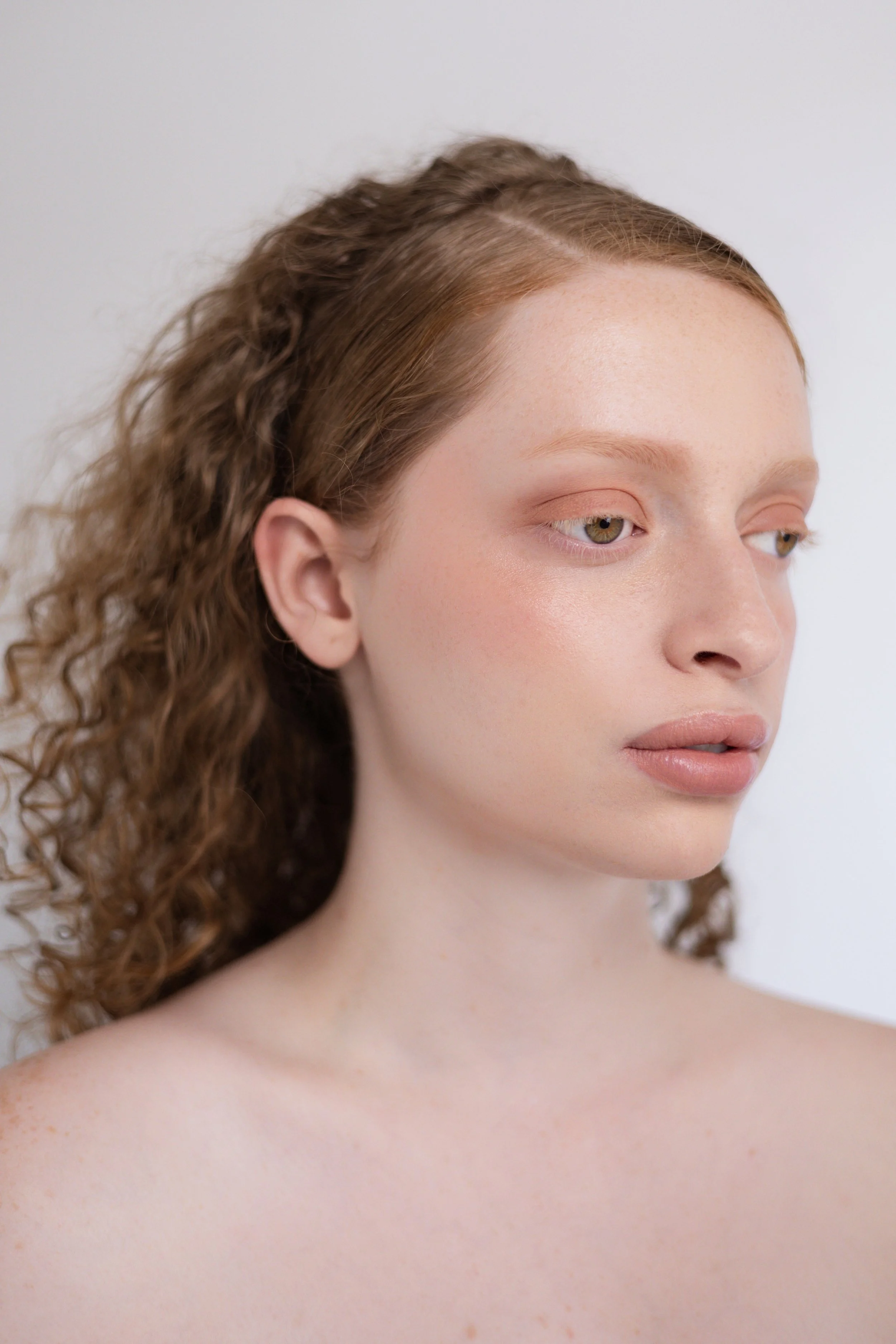 Shooting beauté. Shooting beauté. Jeune femme aux cheveux bruns frisés, regardant vers la droite, avec une peau claire et naturelle, posé contre un fond blanc.
