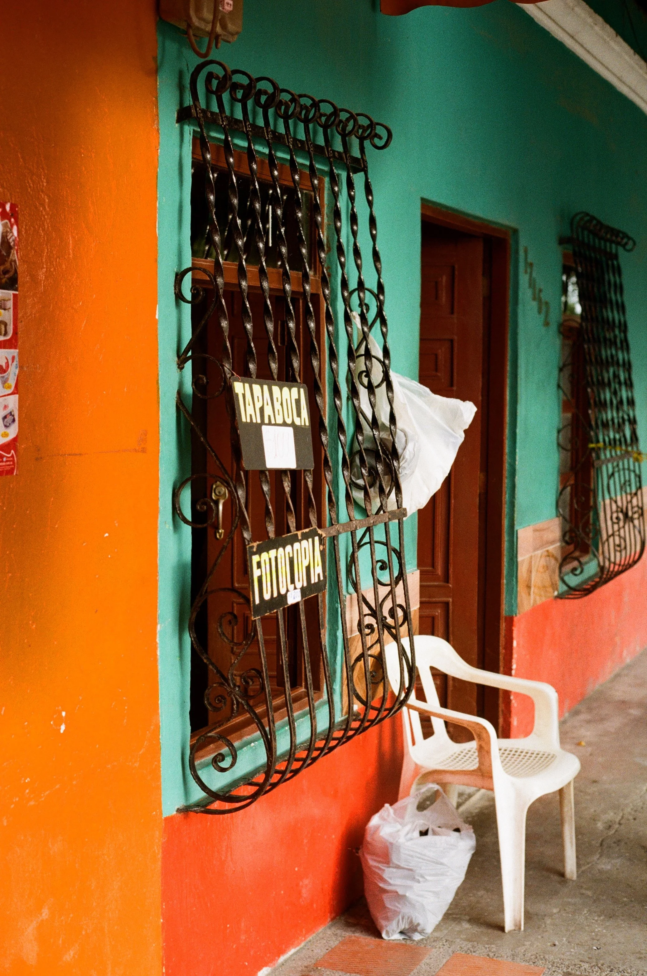 Façade colorée avec fenêtre protégée par une grille en fer forgé, une chaise blanche, un sac en plastique et une porte en bois. Signes indiquant 'TAPABOCA' et 'FOTOCOPIA' suspendus à la grille.