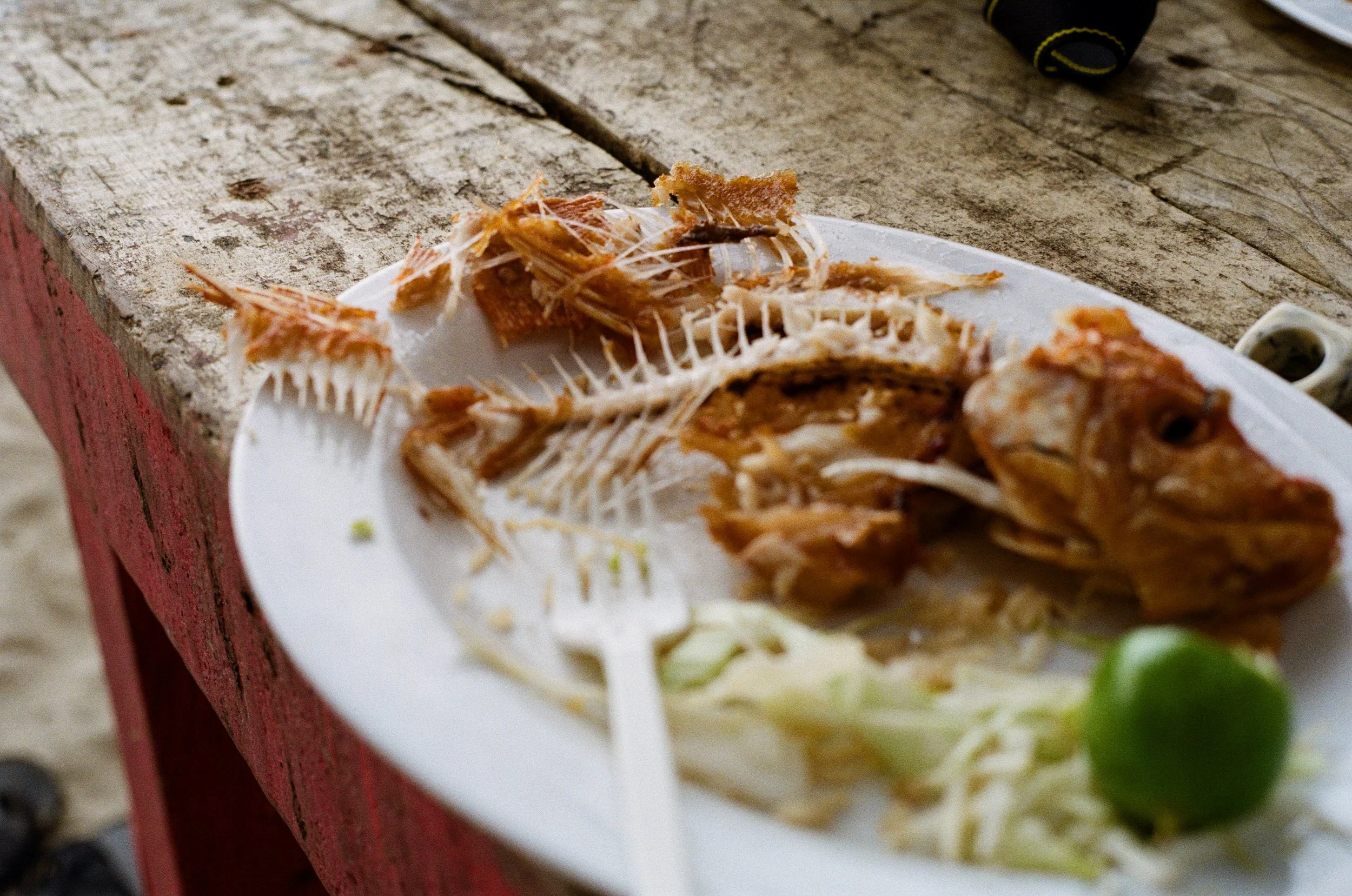 Un poisson entier frit sur une assiette en plastique blanc, avec des restes d'os et de chair, accompagnée de laitue et d'un citron vert, posée sur une table en bois rustique.