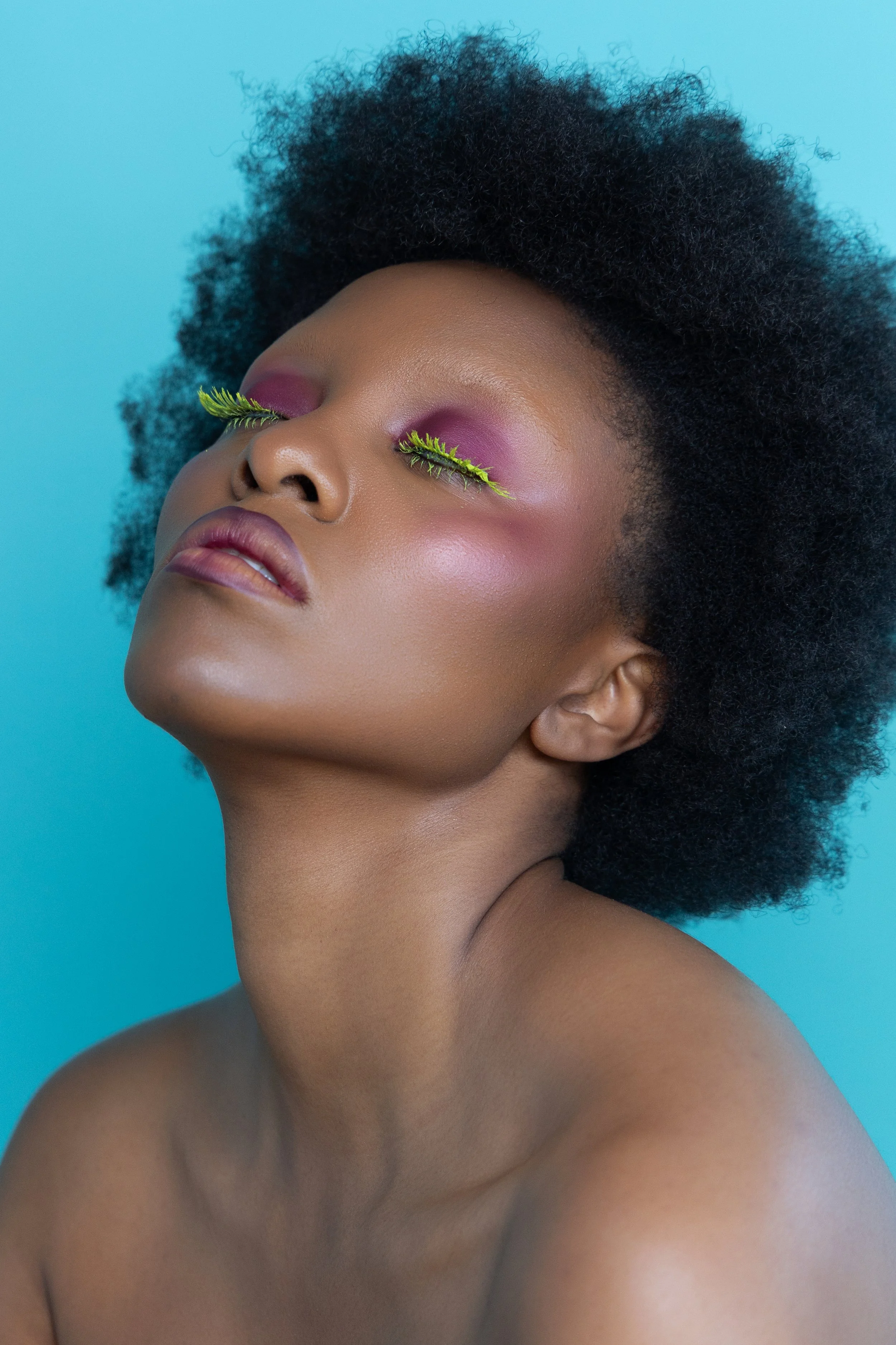 Photo d'une femme avec des extensions de cils faites avec des feuilles vertes, portant un maquillage violet et un fond bleu.