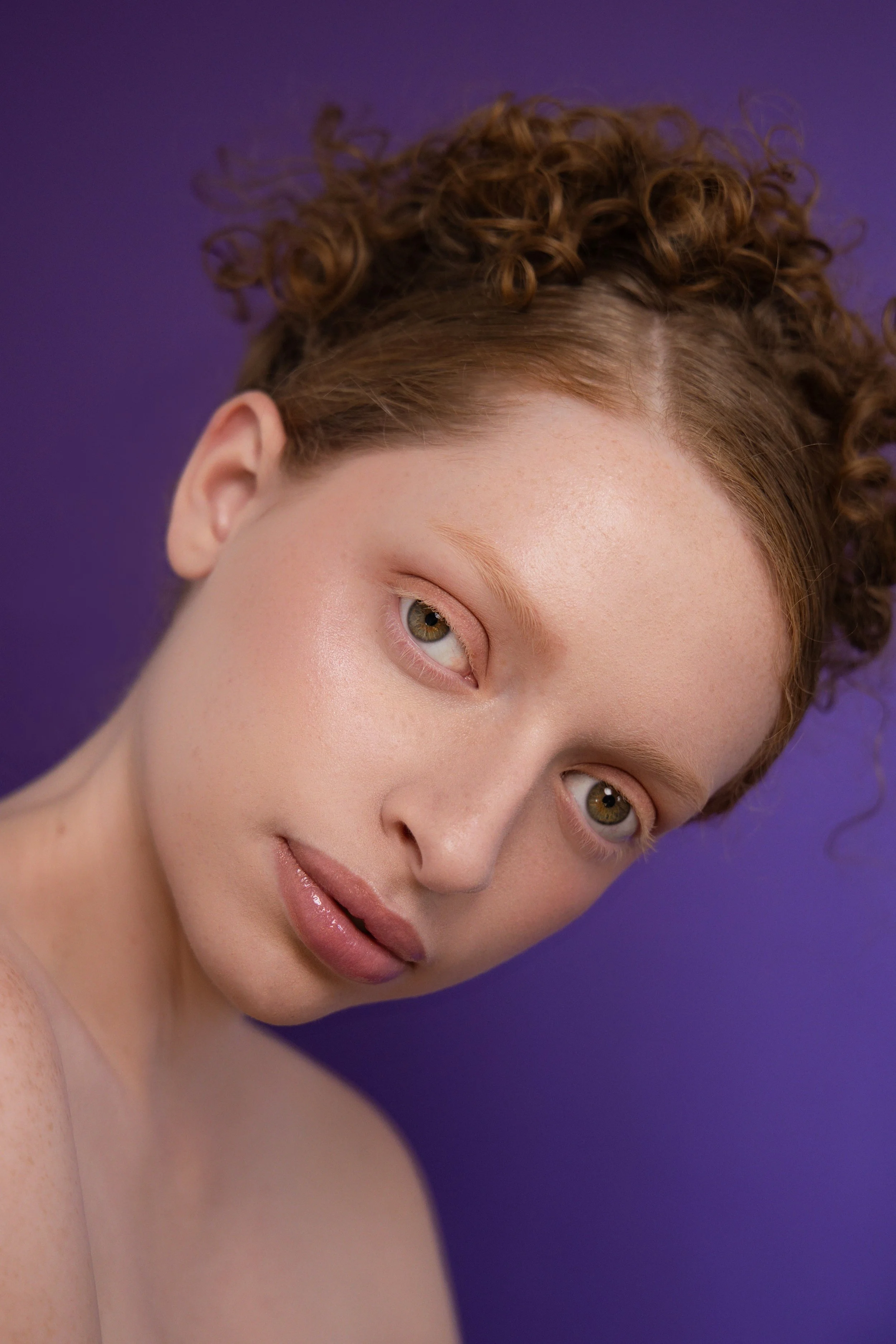 Shooting beauté. Portrait d'une jeune femme avec des cheveux bruns bouclés, peau claire, et regardant légèrement vers la gauche, positionnée de profil contre un fond violette