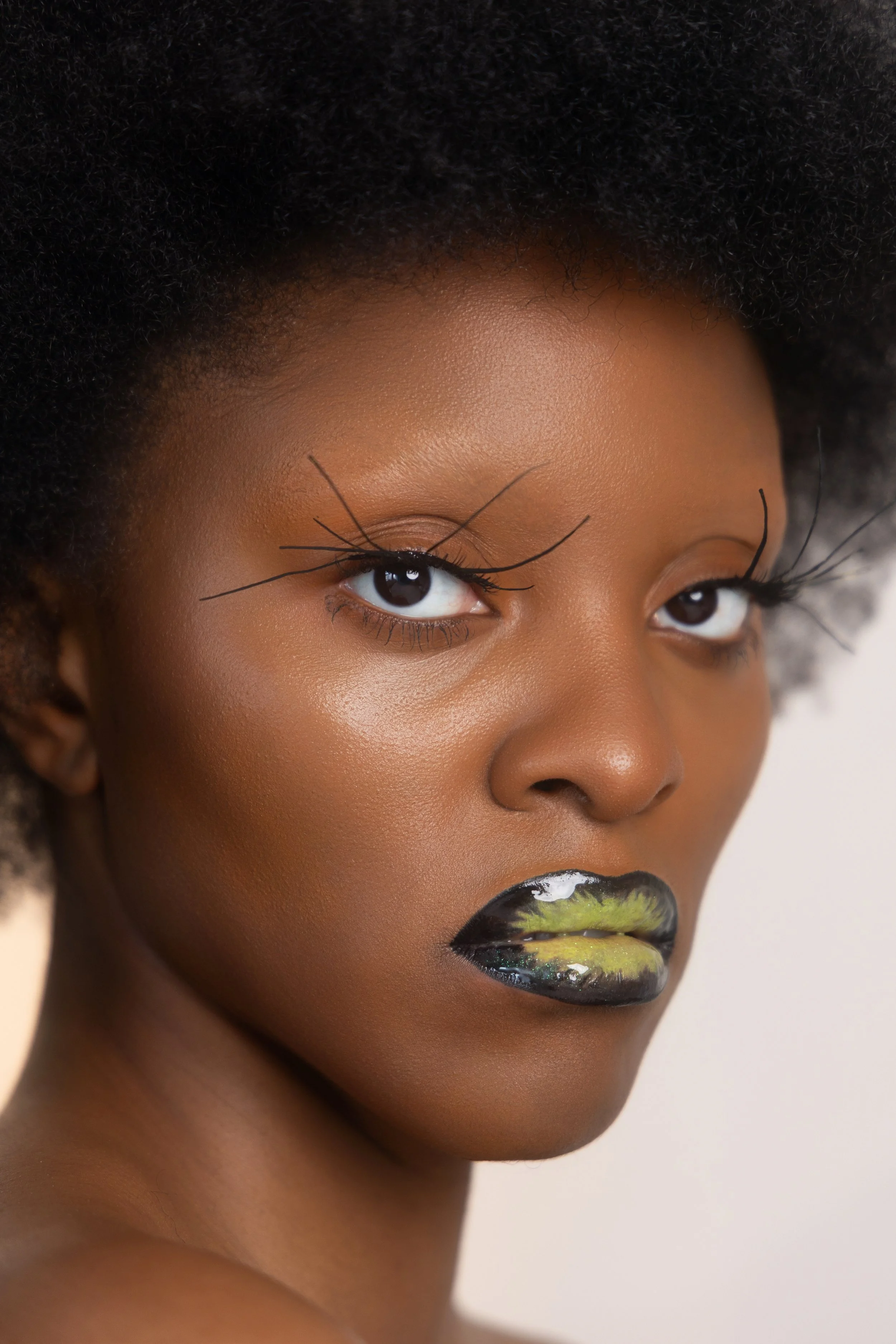 Shooting beauté. Portrait d'une femme noire avec un maquillage artistique, notamment des lèvres peintes en noir avec des touches de vert, et des longs cils apparemment artificiels ou très accentués.