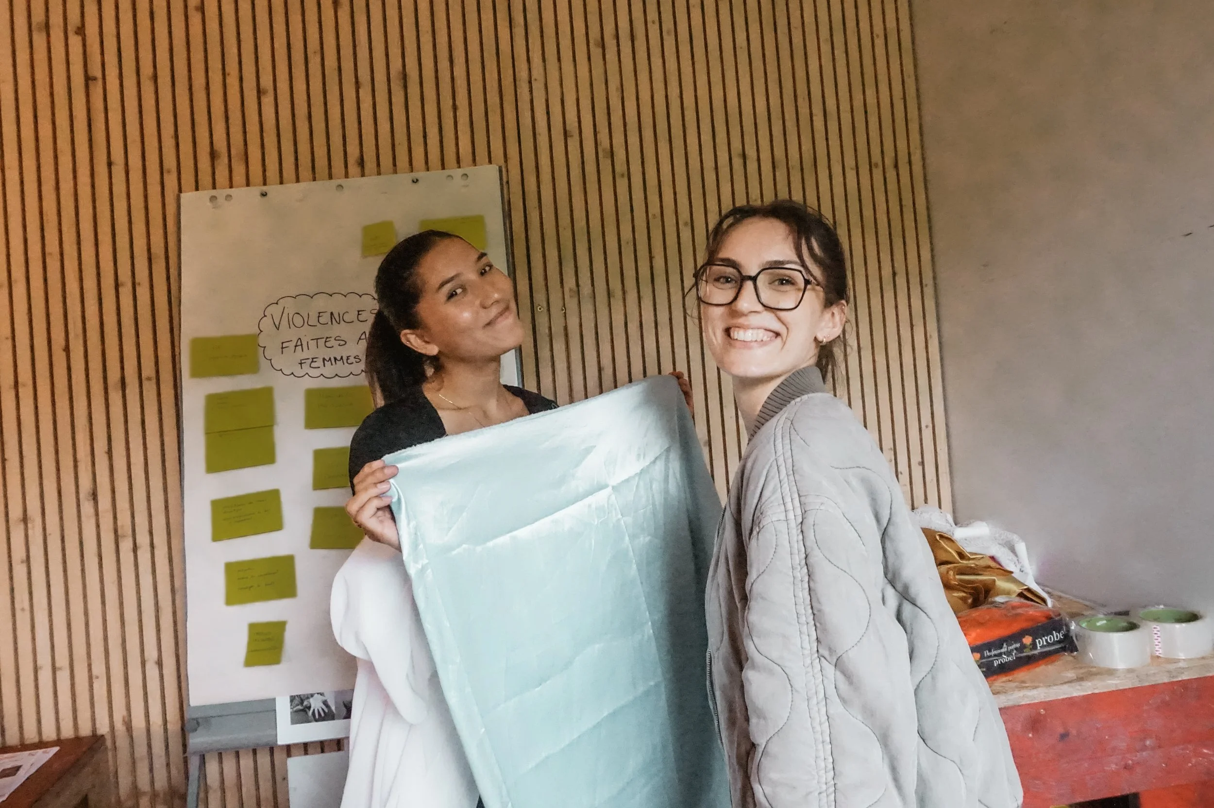 Deux jeunes femmes souriantes qui tiennent une boîte, dans une salle avec un fond en bois et un tableau à post-it avec le texte "Violence faites aux femmes".