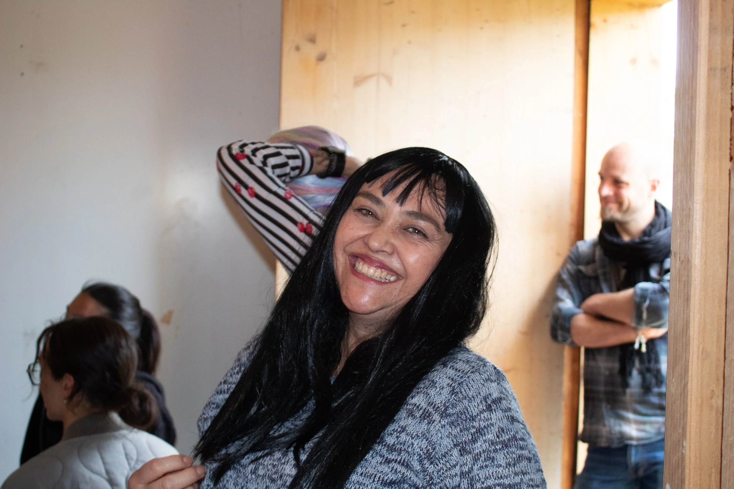 Une femme souriante avec des cheveux noirs longs, portant un vêtement gris à motifs, dans une pièce avec des autres personnes en arrière-plan.