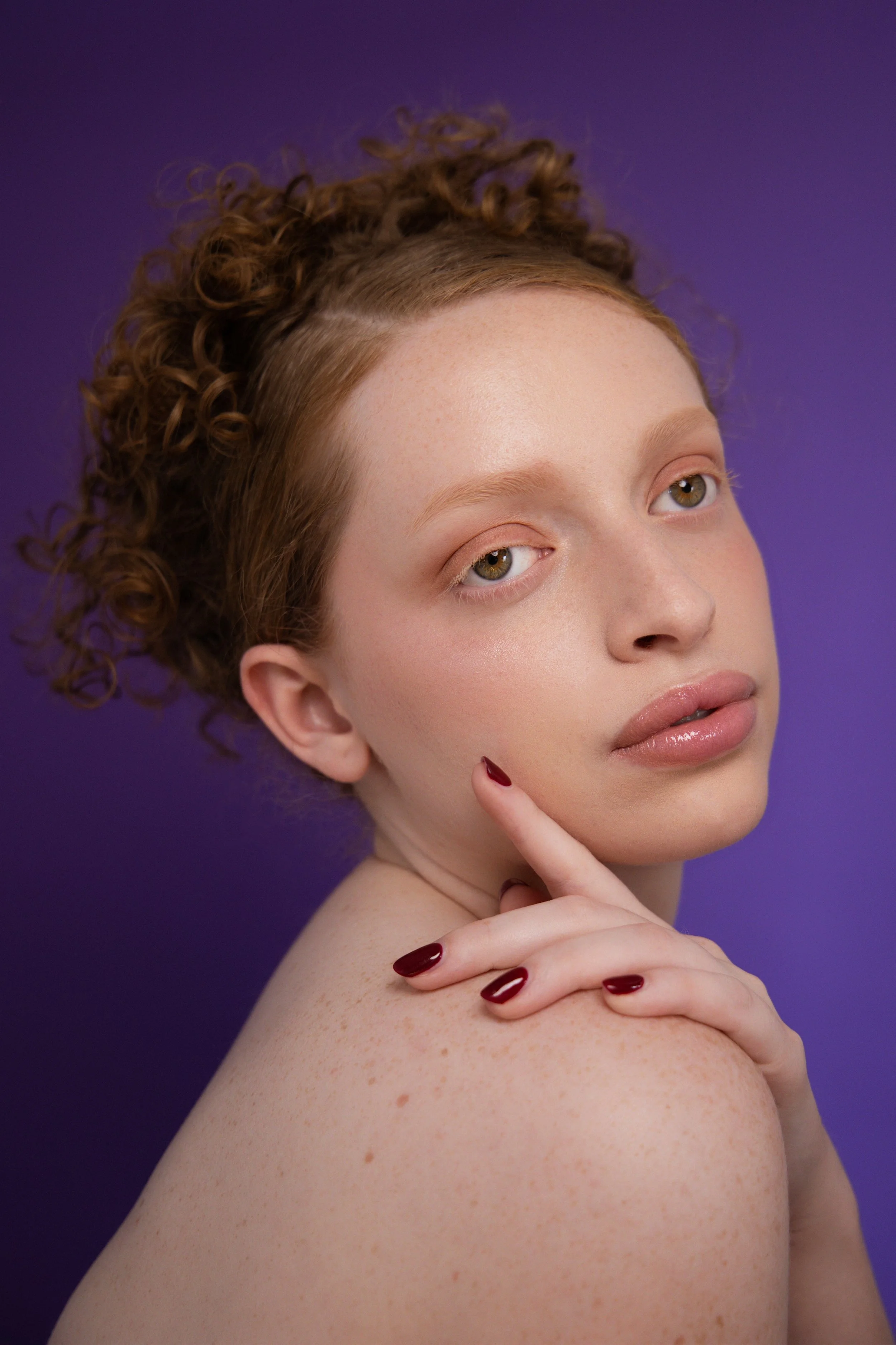 Shooting beauté. Portrait d'une femme aux cheveux roux bouclés, regardant vers l'objectif, avec une main sur l'épaule, fond violet.