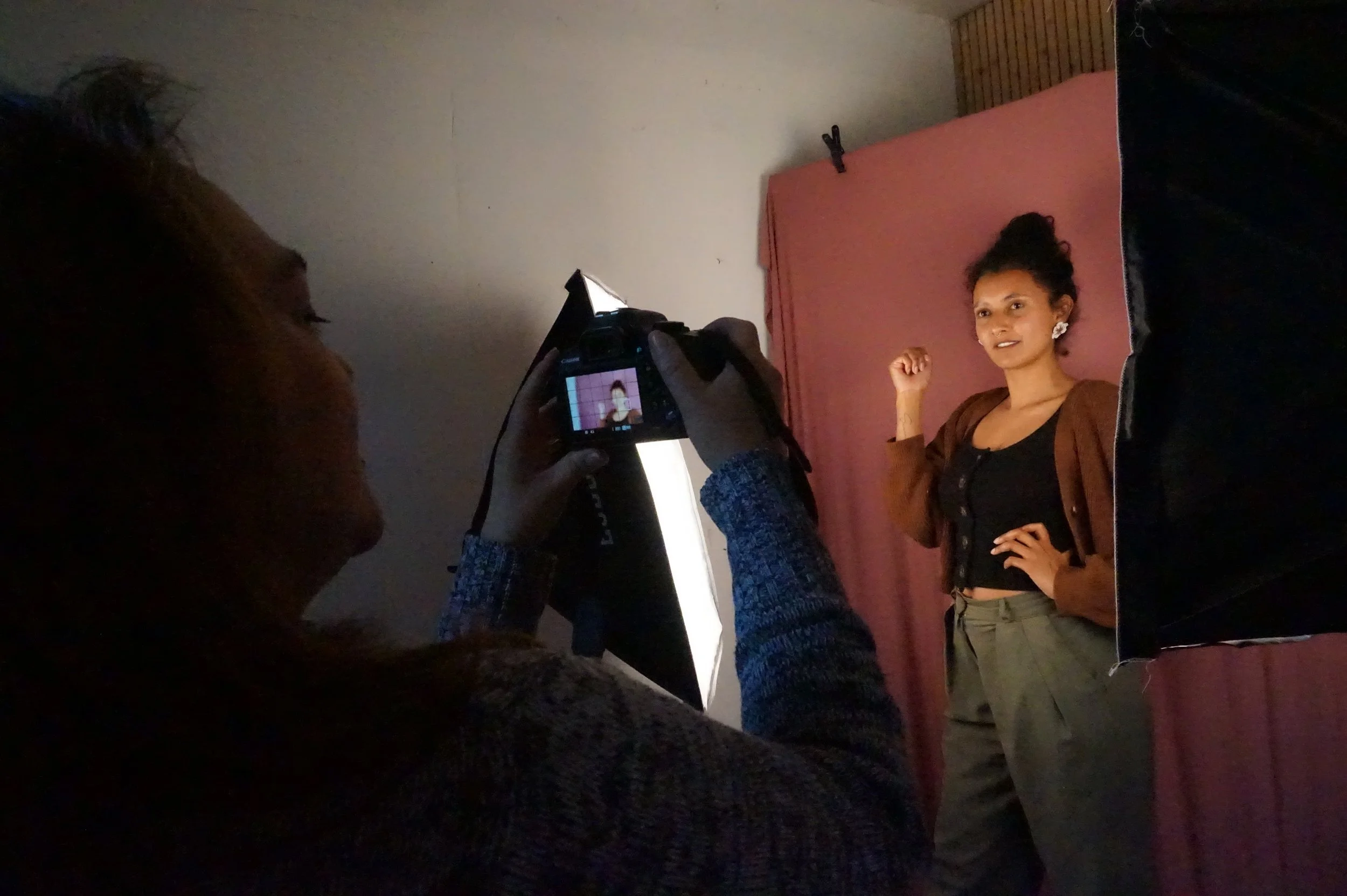 Une personne photographie une femme posant devant un fond rose, dans un studio de photographie, utilisant un appareil photo numérique.