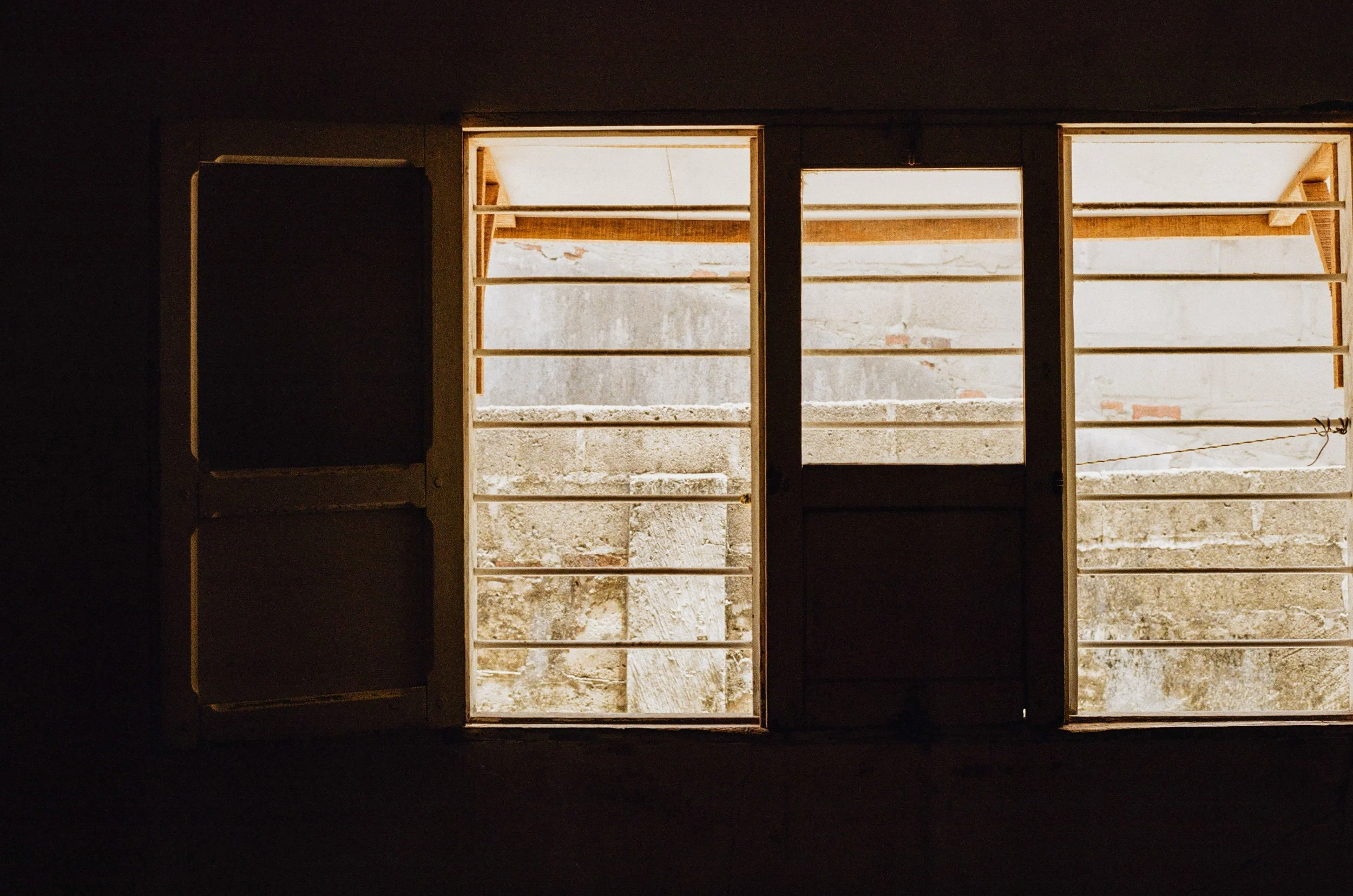 Fenêtres en bois laissant passer la lumière, vue de l'intérieur, donnant sur un mur en pierre ou béton.