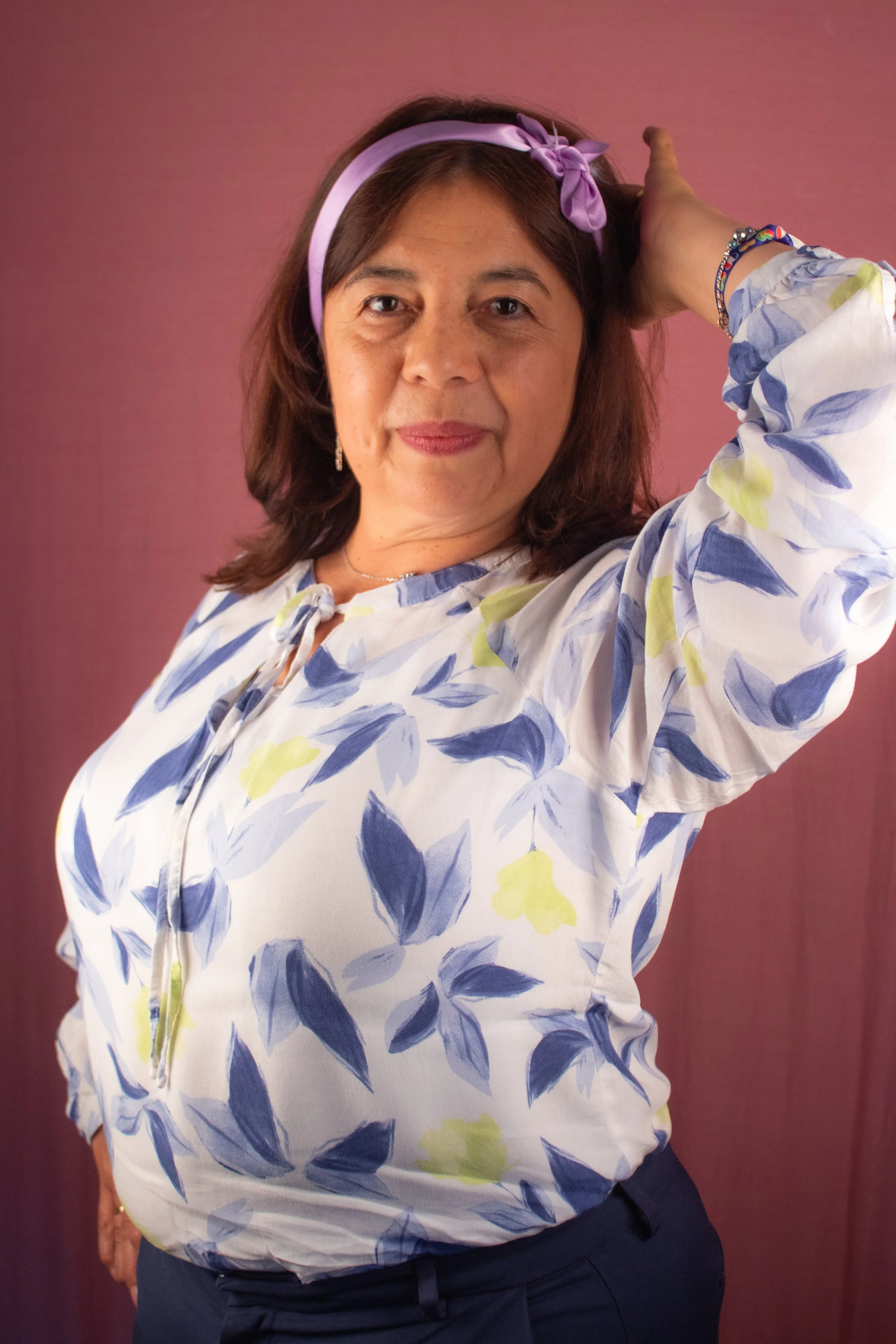 Une femme posant devant un fond rose, portant une chemise blanche à motifs floraux bleus et verts, une bandeau violet dans les cheveux, elle regarde l'objectif avec un léger sourire.