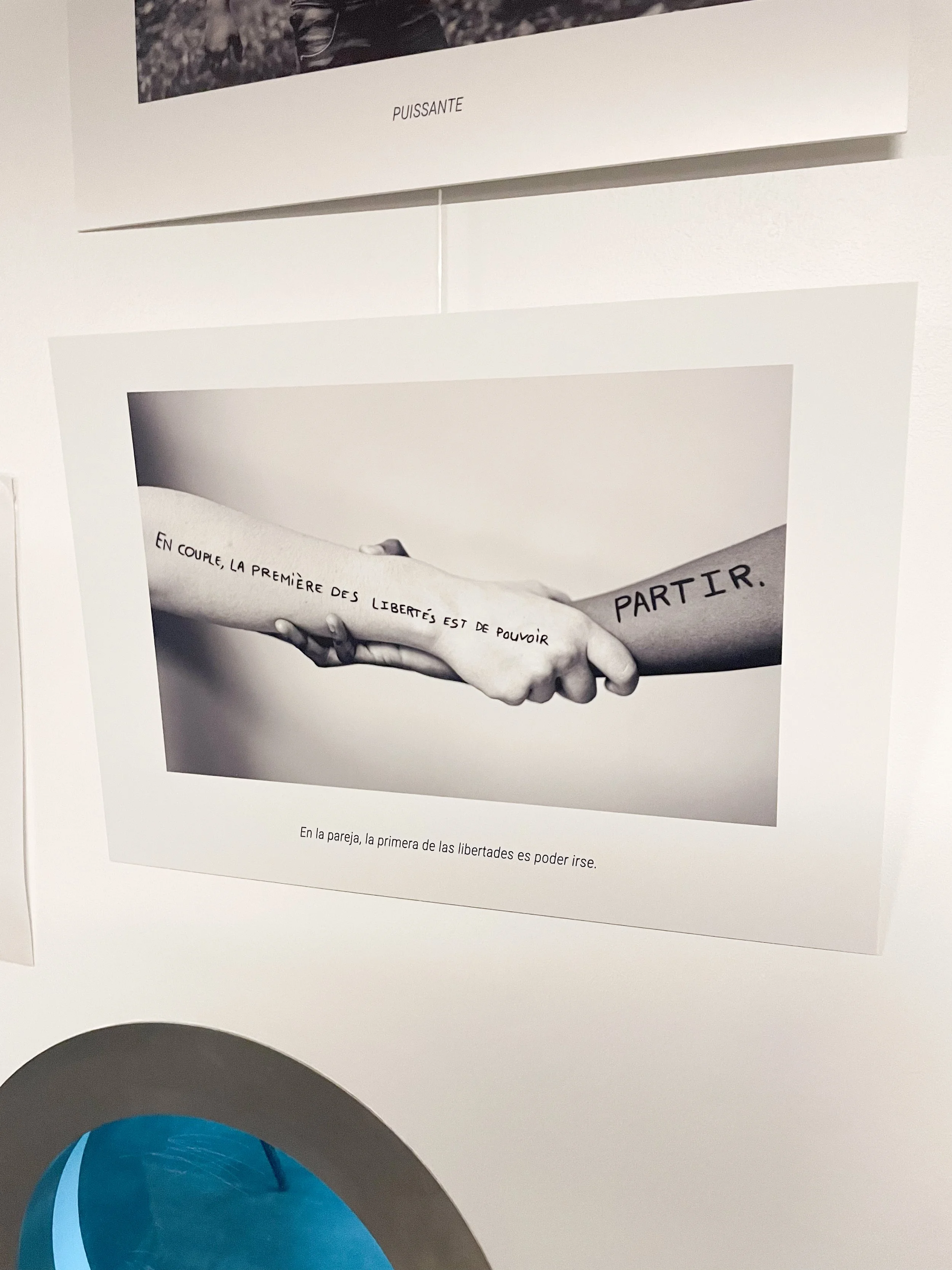 Photographie en noir et blanc d'une poignée de main entre deux personnes, avec des tatouages sur leur bras. Le tatouage sur le bras gauche dit "EN COUPLE, LA PREMIÈRE DES LIBERTÉS EST DE POUVOIR" et celui sur le bras droit dit "PARTIR".