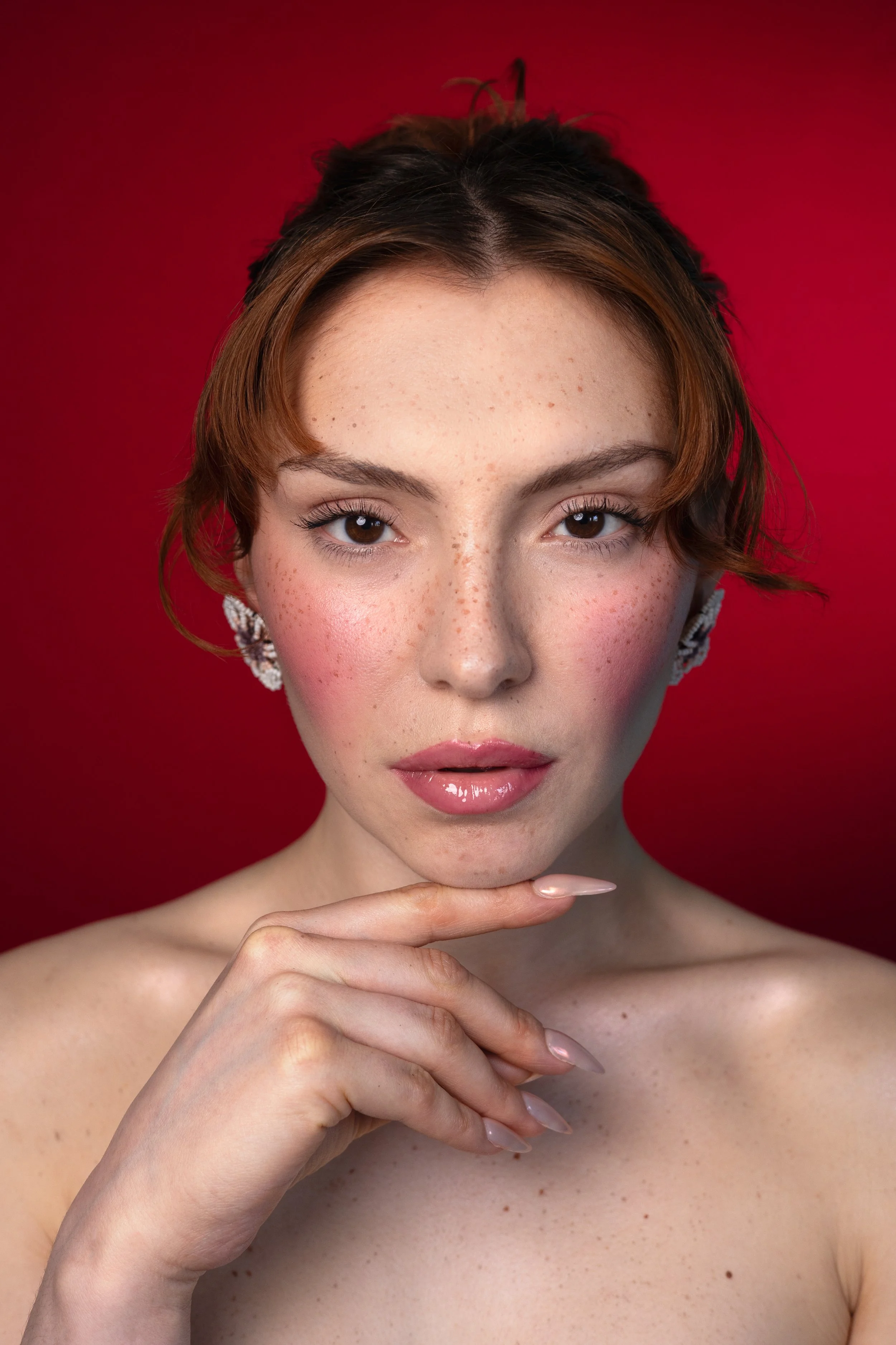 Portrait d'une jeune femme avec des taches de rousseur, portant des boucles d'oreilles, contre un fond rouge, avec une expression confiante, touchant doucement son menton.