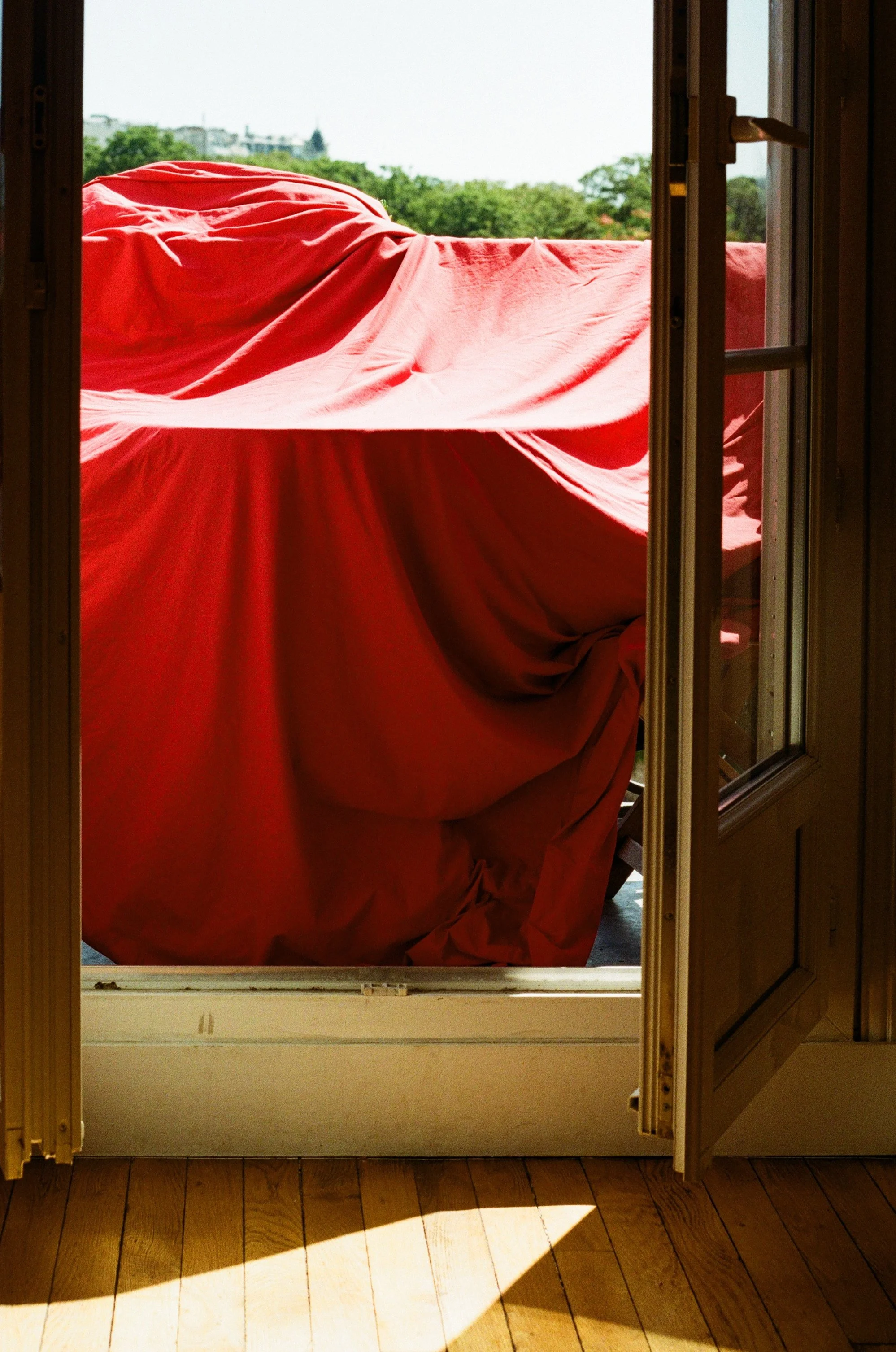 Tableau d'une porte-fenêtre ouverte donnant sur une terrasse avec une draperie rouge suspendue, vue sur des arbres et un ciel clair.