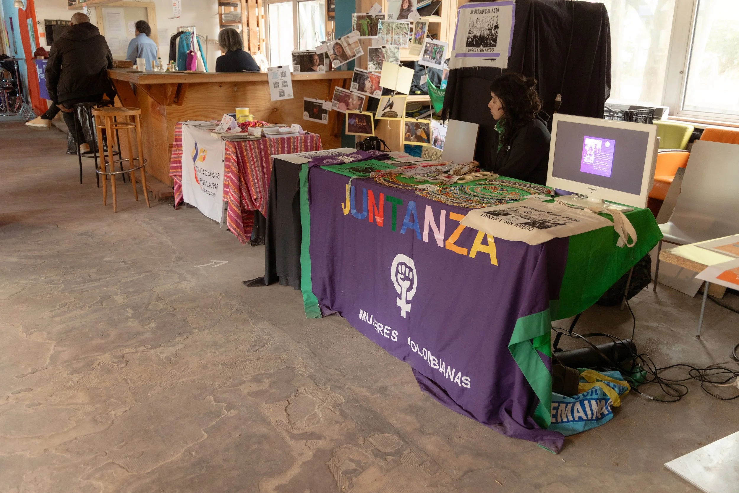 Stand avec un drapeau violet, vert et noir affichant 'JUNTANZA MUJERES COLOMBIANAS' et une femme assise devant un ordinateur, des bijoux ou accessoires sur la table.