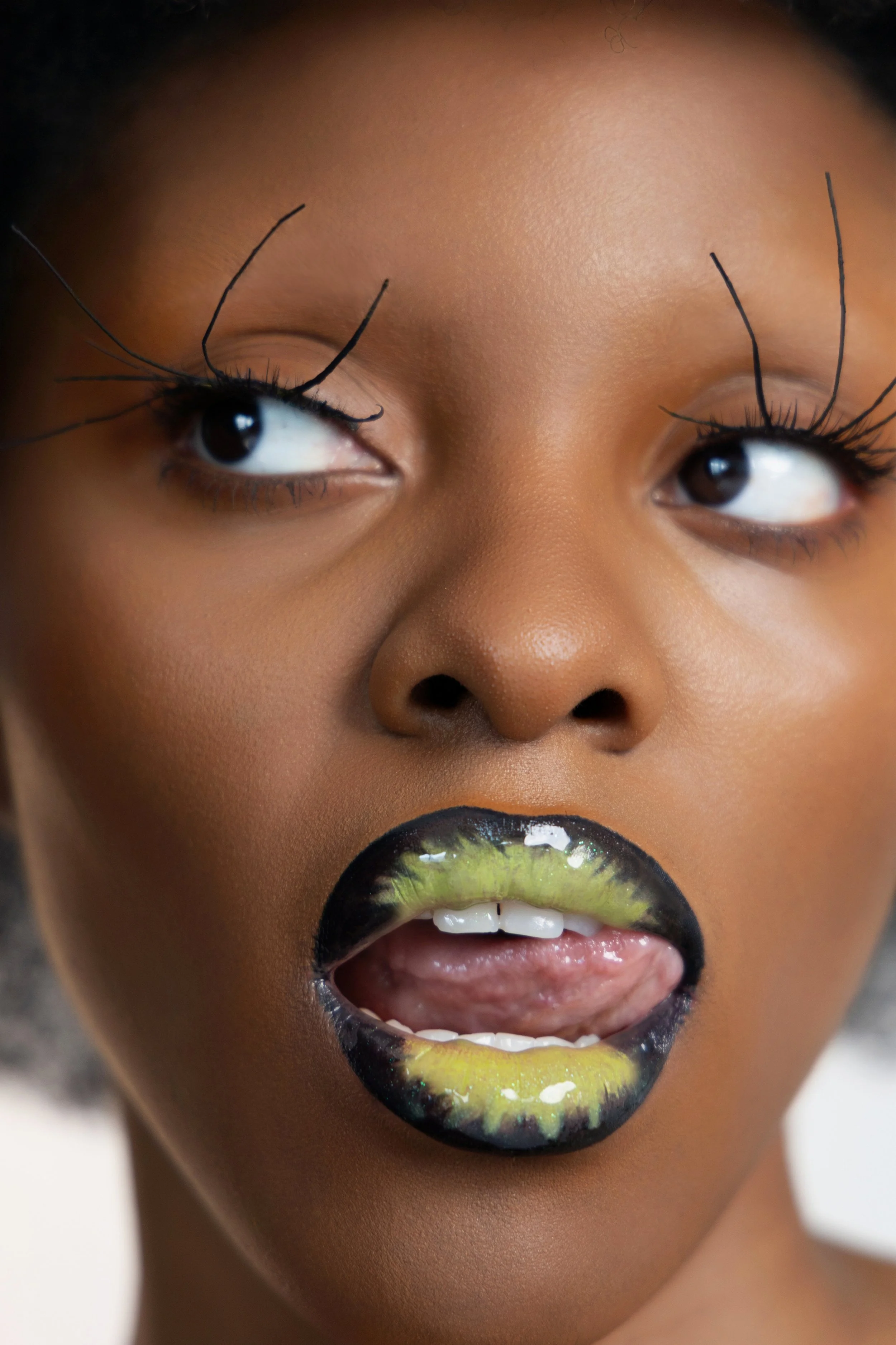 Portrait d'une femme noire avec maquillage artistique, bavant et regardant sur le côté, avec des cils longs et des lèvres noires et vertes.