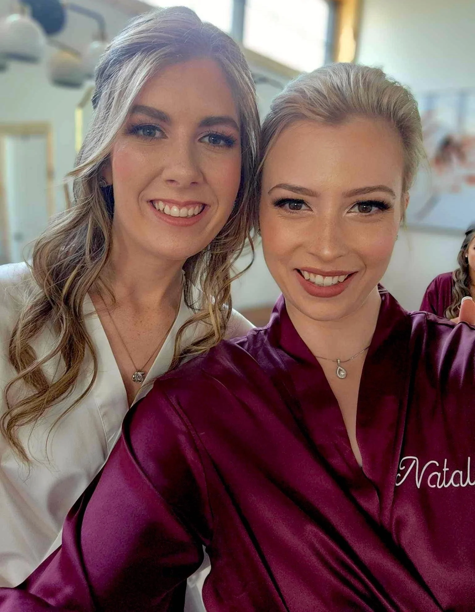 Two women in robes smiling and posing for a selfie, showcasing their makeup as the bride and bridesmaid get ready for the big day