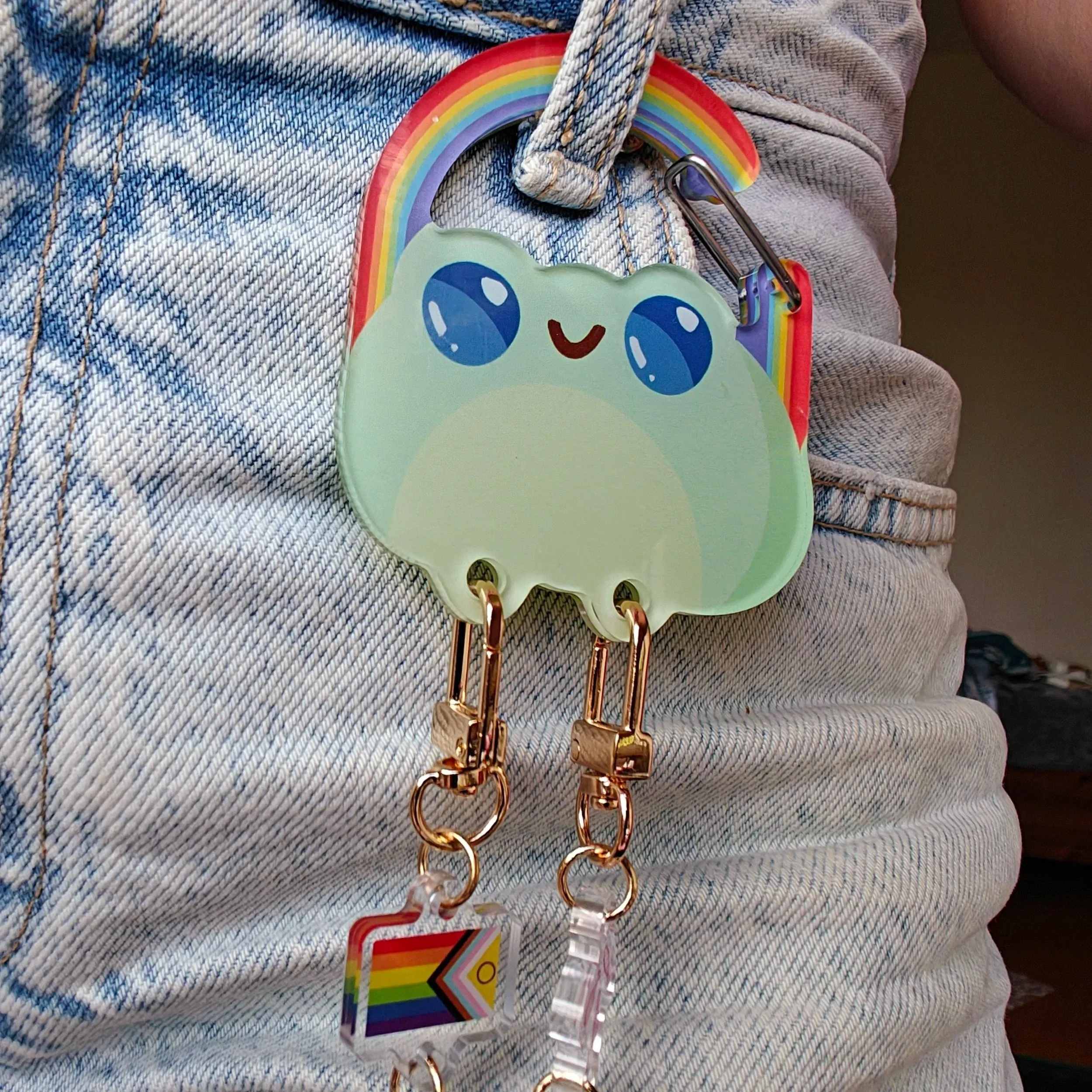 Close-up of a rainbow, frog-shaped carabiner with a smiling face, attached to a belt loop on light-colored denim jeans. The holder has two clips at the bottom with a rainbow flag charm attached.