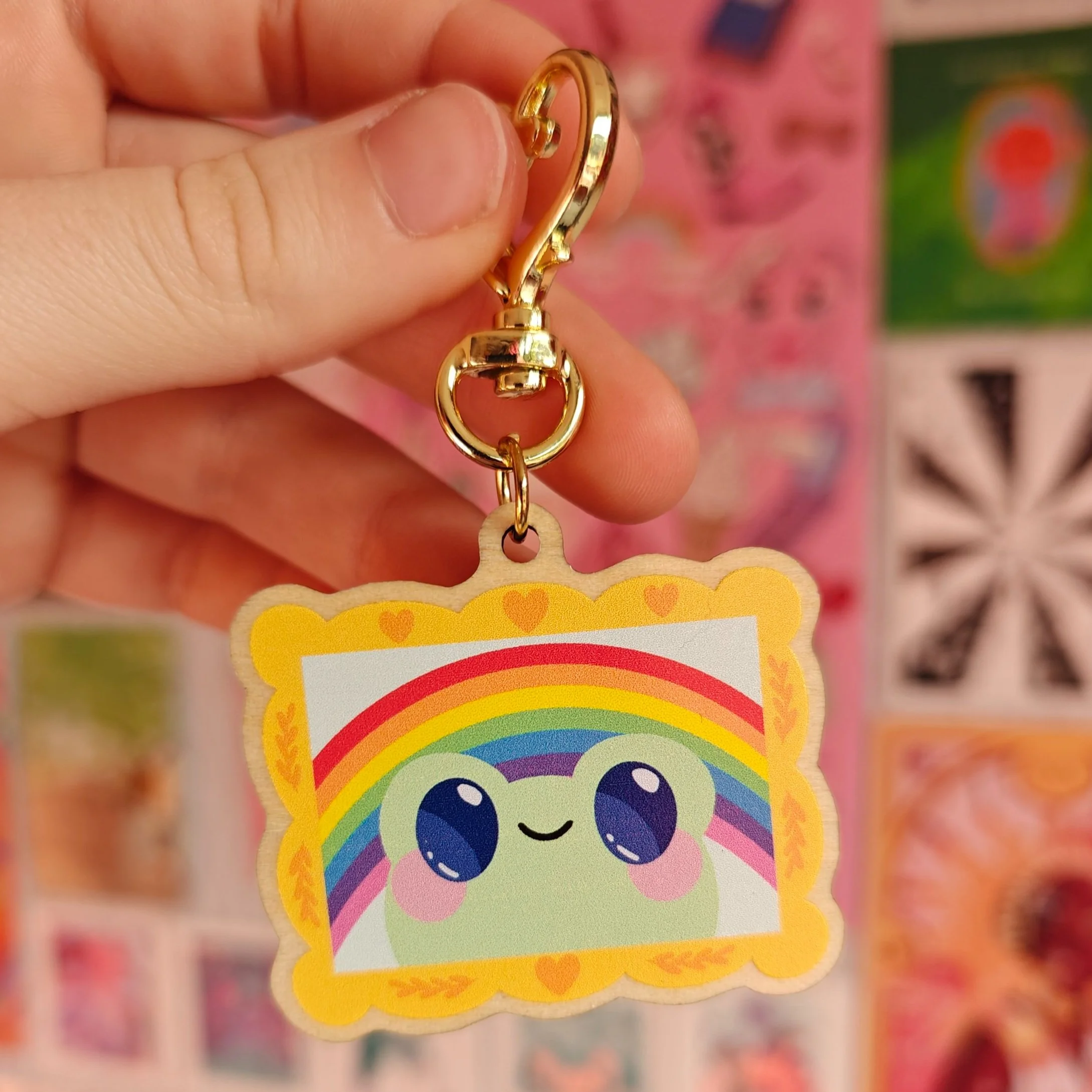 A keychain featuring a cute, smiling cartoon character with big eyes, set against a rainbow background and surrounded by a yellow border with small hearts.