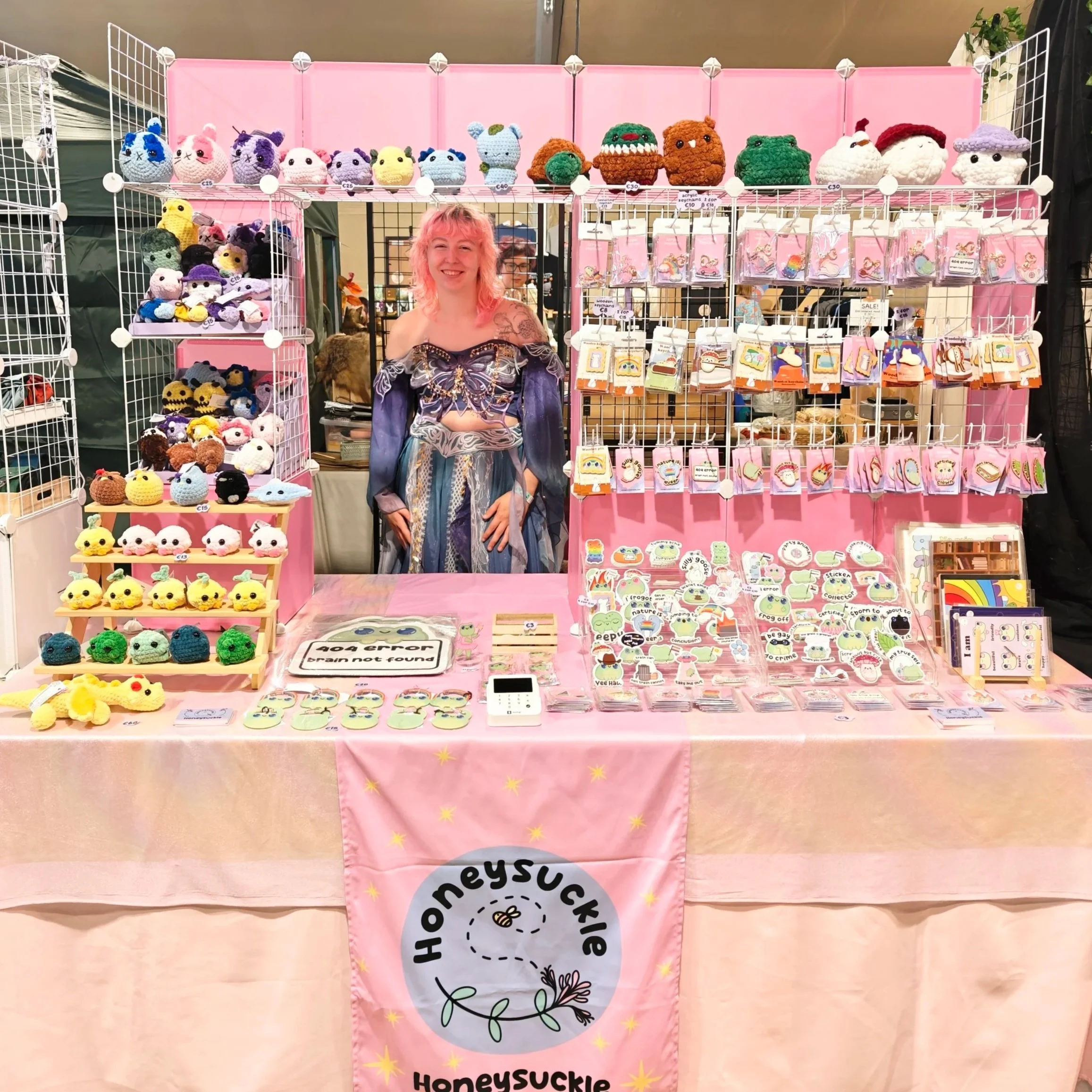 A person with pink hair standing behind a pink display table at Elfia Arcen, showcasing various handmade plush toys, patches, and stickers, with a pink Honeysuckle banner hanging from the front of the table.