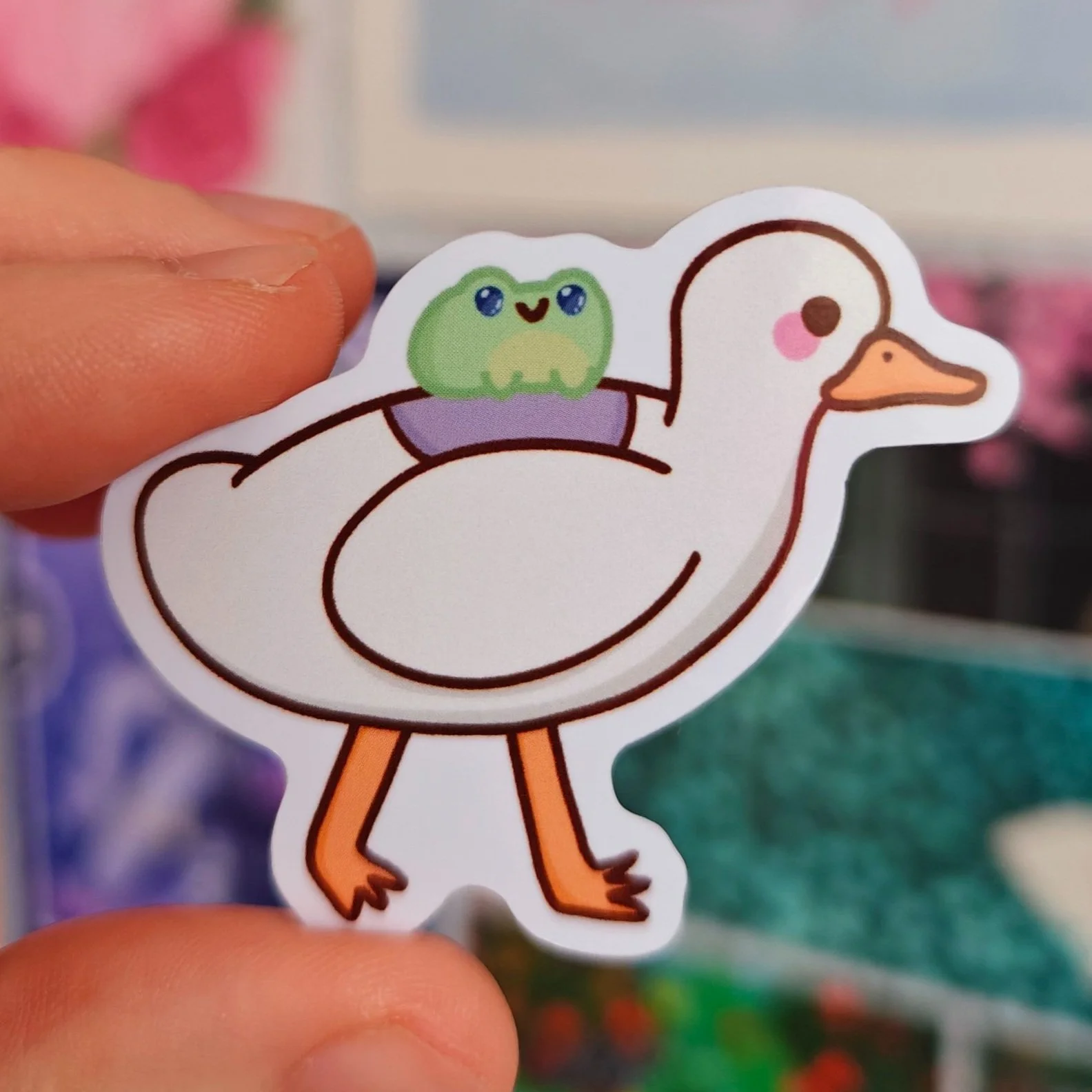 A sticker of a cartoon goose with an orange beak and legs, having a small green frog with big blue eyes sitting on its back.
