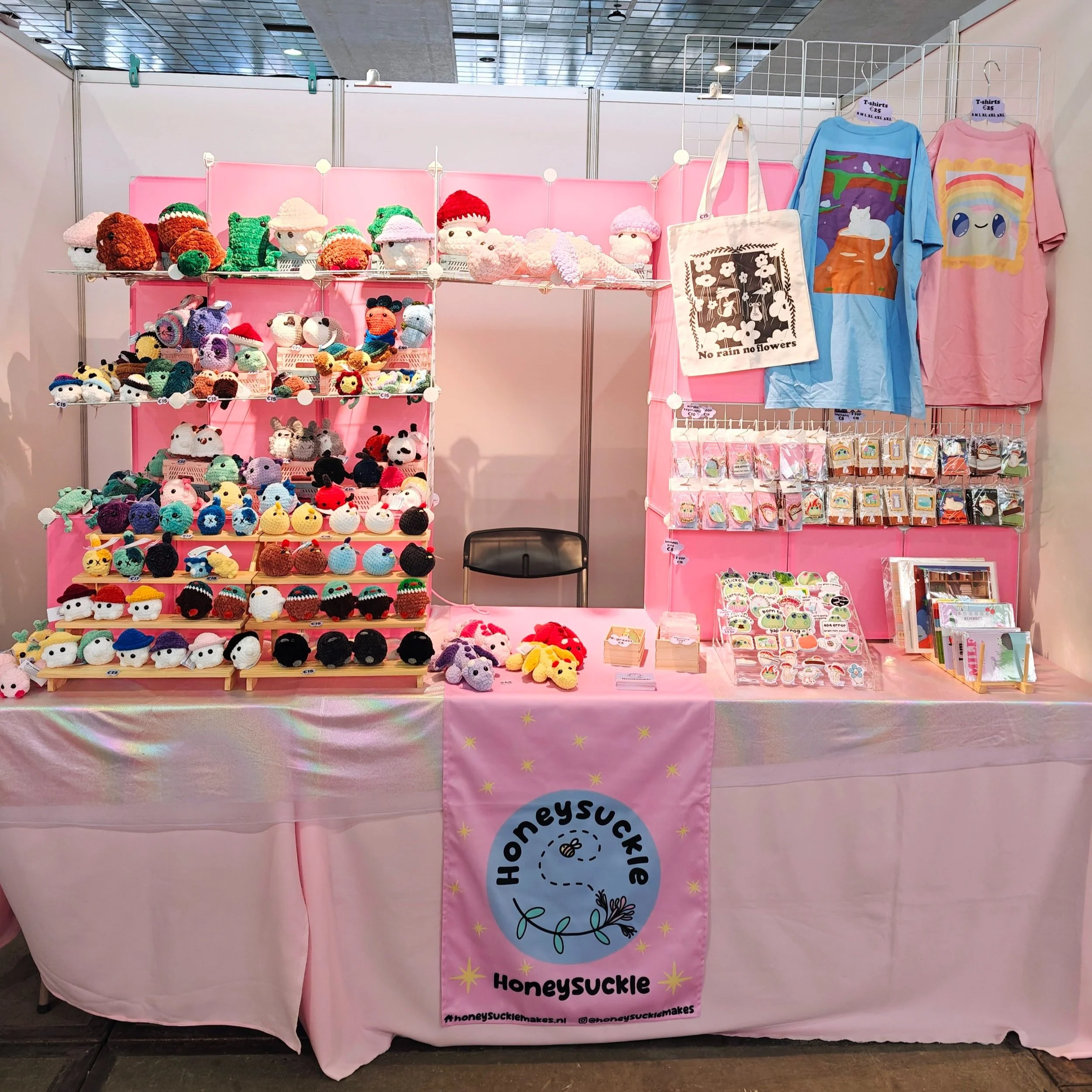 A booth selling handmade crochet plush toys, tote bags, stickers, and notebooks at Made In Asia Brussels. The booth has a pink backdrop and pink tablecloth with a banner that reads 'Honeysuckle'.