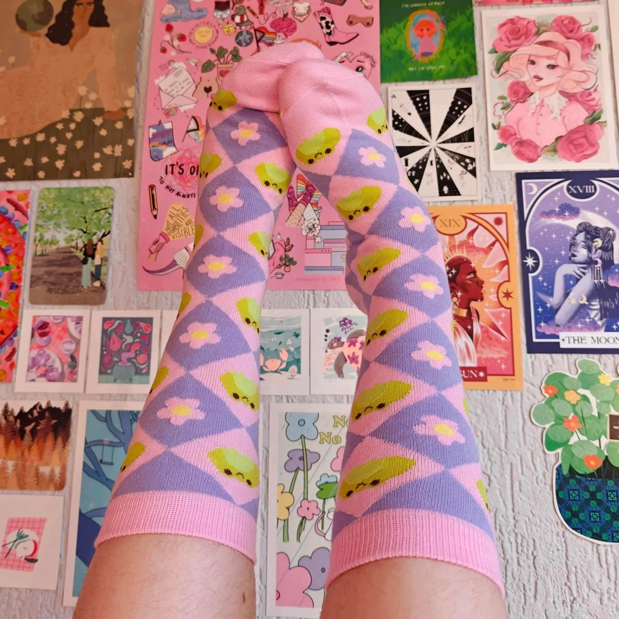 A pair of legs wearing pastel pink socks with a pattern of green frog faces and pink flowers, extended upward in front of a wall decorated with colorful illustrated postcards and posters featuring various artistic and whimsical designs.