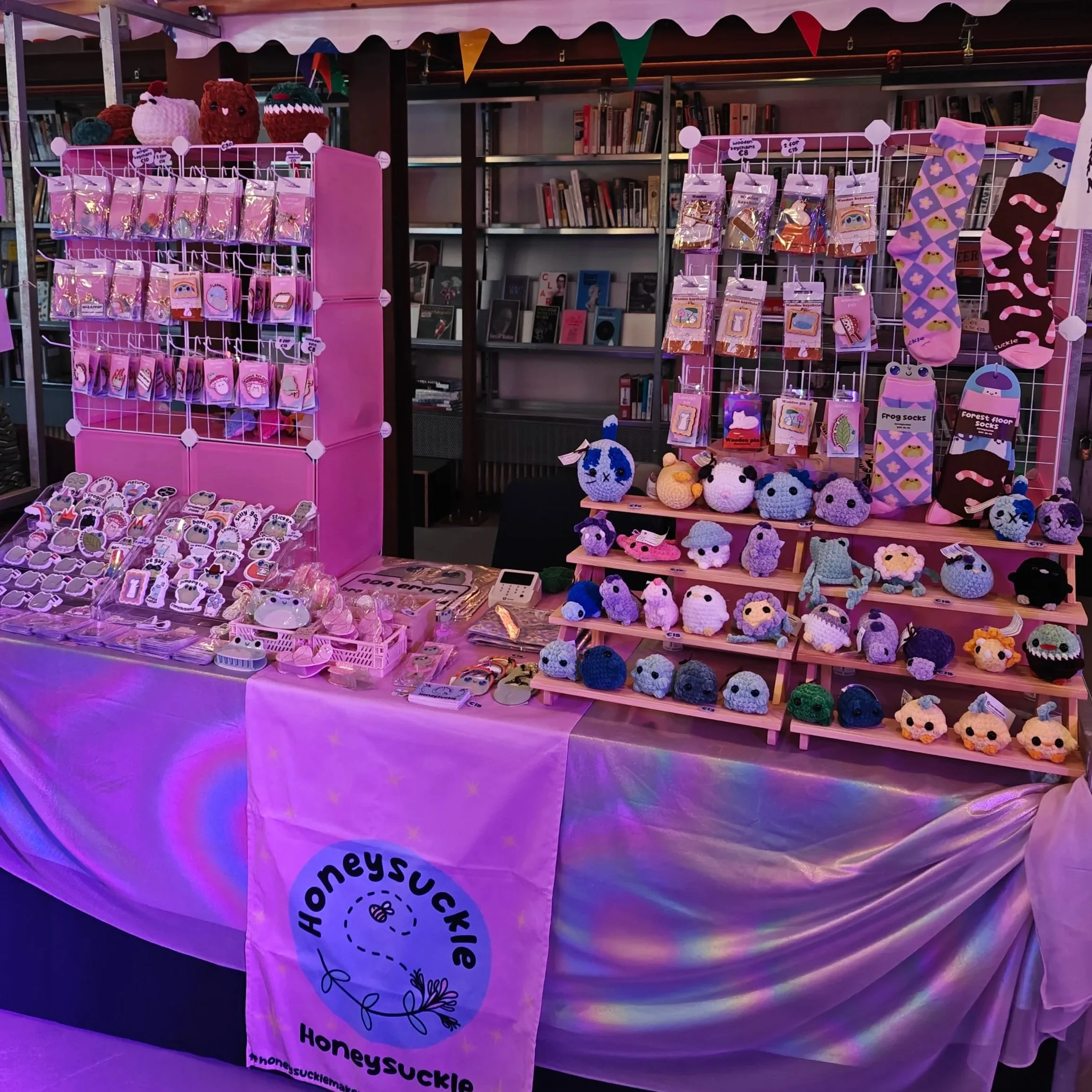 A pink display booth featuring handmade plush toys, keychains, socks, and small accessories at Creative CoLab in Breda. The booth features cute frog art and quirky designs.