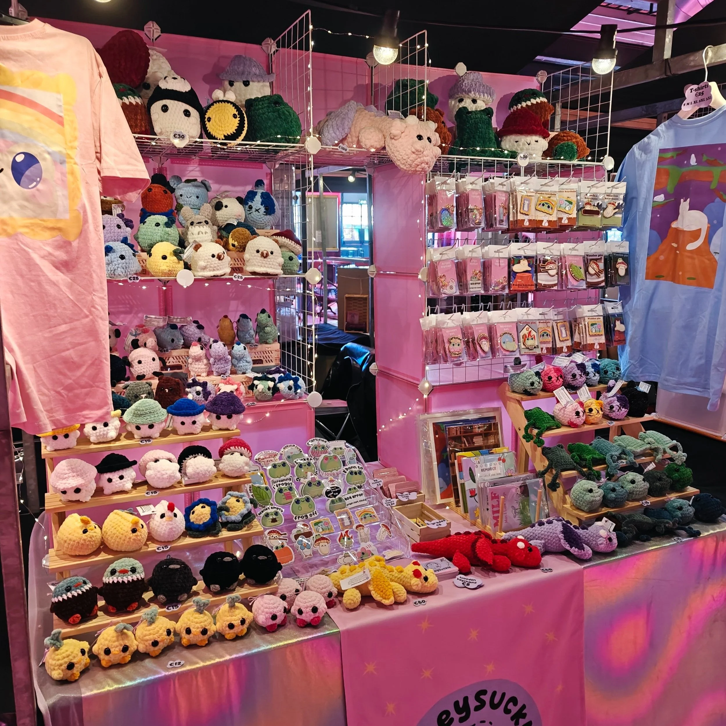 A display booth at Playgrounds TAD selling various handmade crocheted plush toys, pins, and stationery with a pink, iridescent tablecloth and pink background. The plush toys include animals and cartoon characters in different colors and sizes.