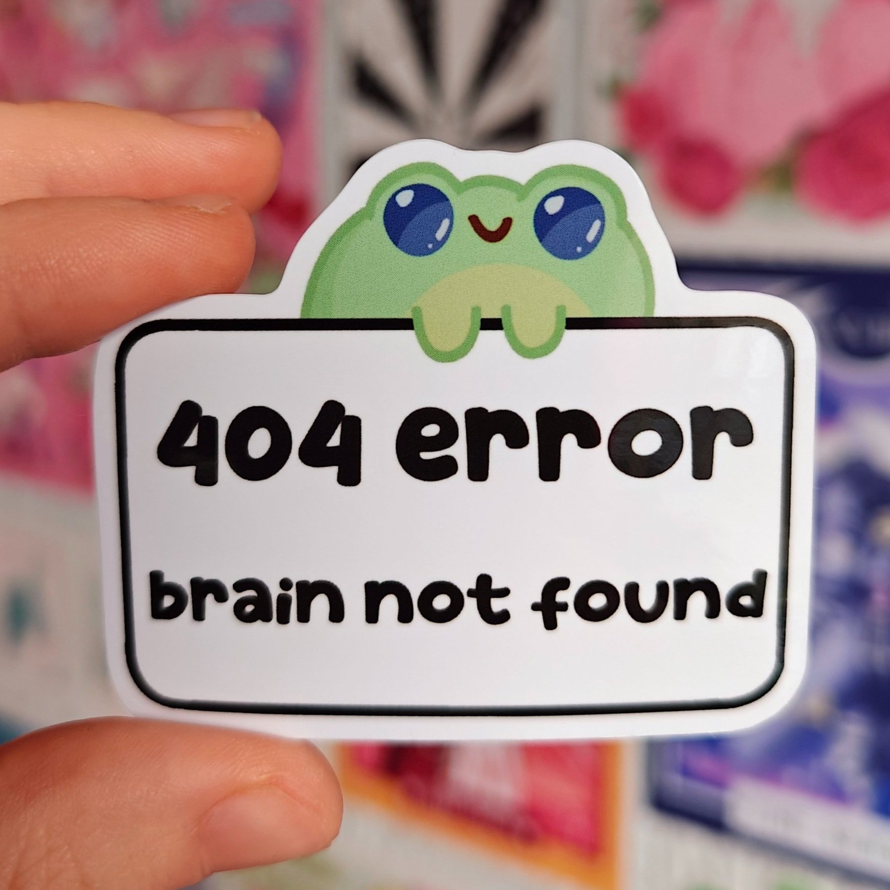 Sticker of a cute green cartoon frog with big blue eyes and a happy face, holding a sign that reads '404 error brain not found'.