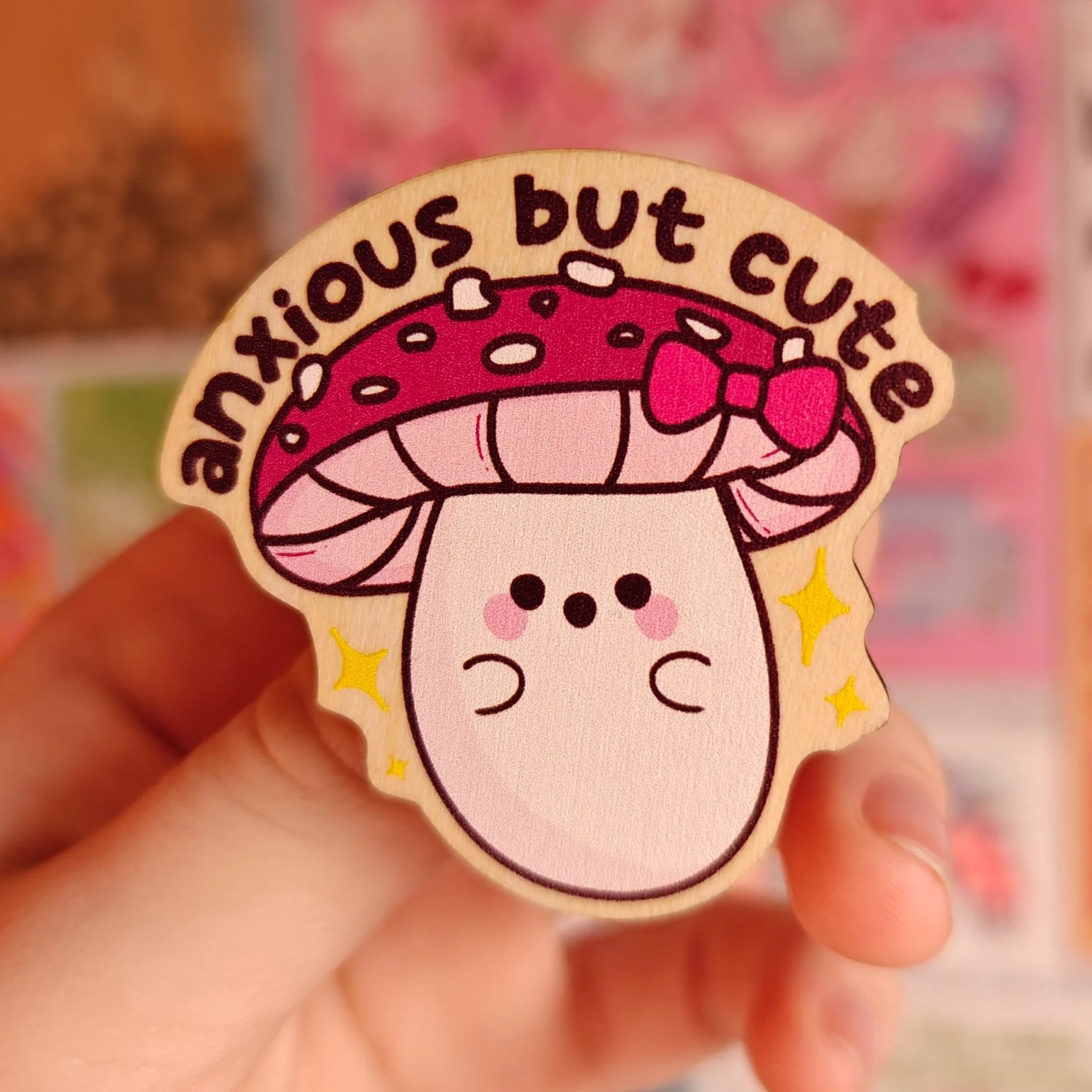A cartoon wooden pin of a cute mushroom with a pink cap and a smiling face, with the words "anxious but cute" written above it, surrounded by small yellow stars.