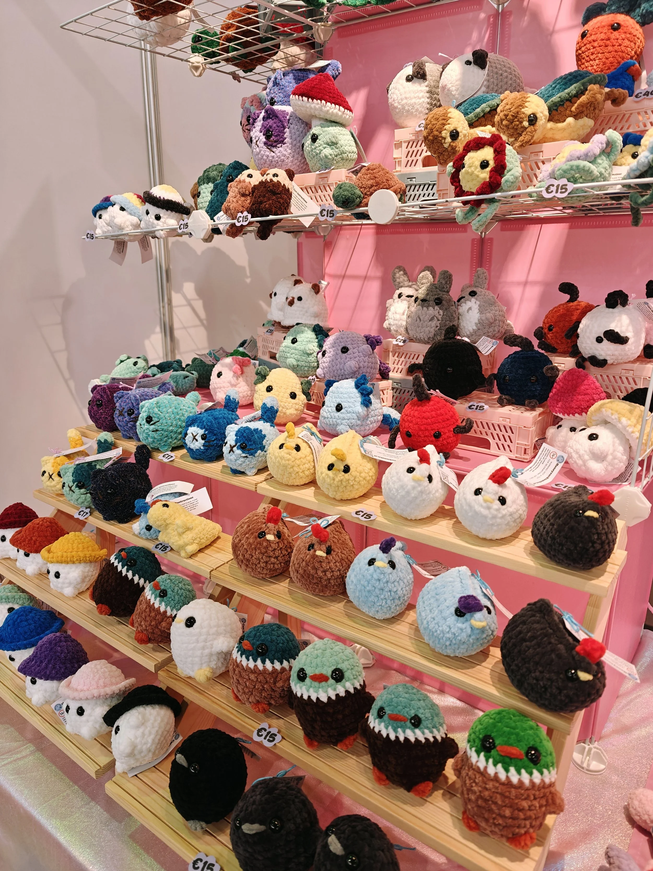 A display of various colorful crocheted animal stuffed toys, including birds, bears, foxes, cats, and other creatures, arranged on wooden and pink shelves.