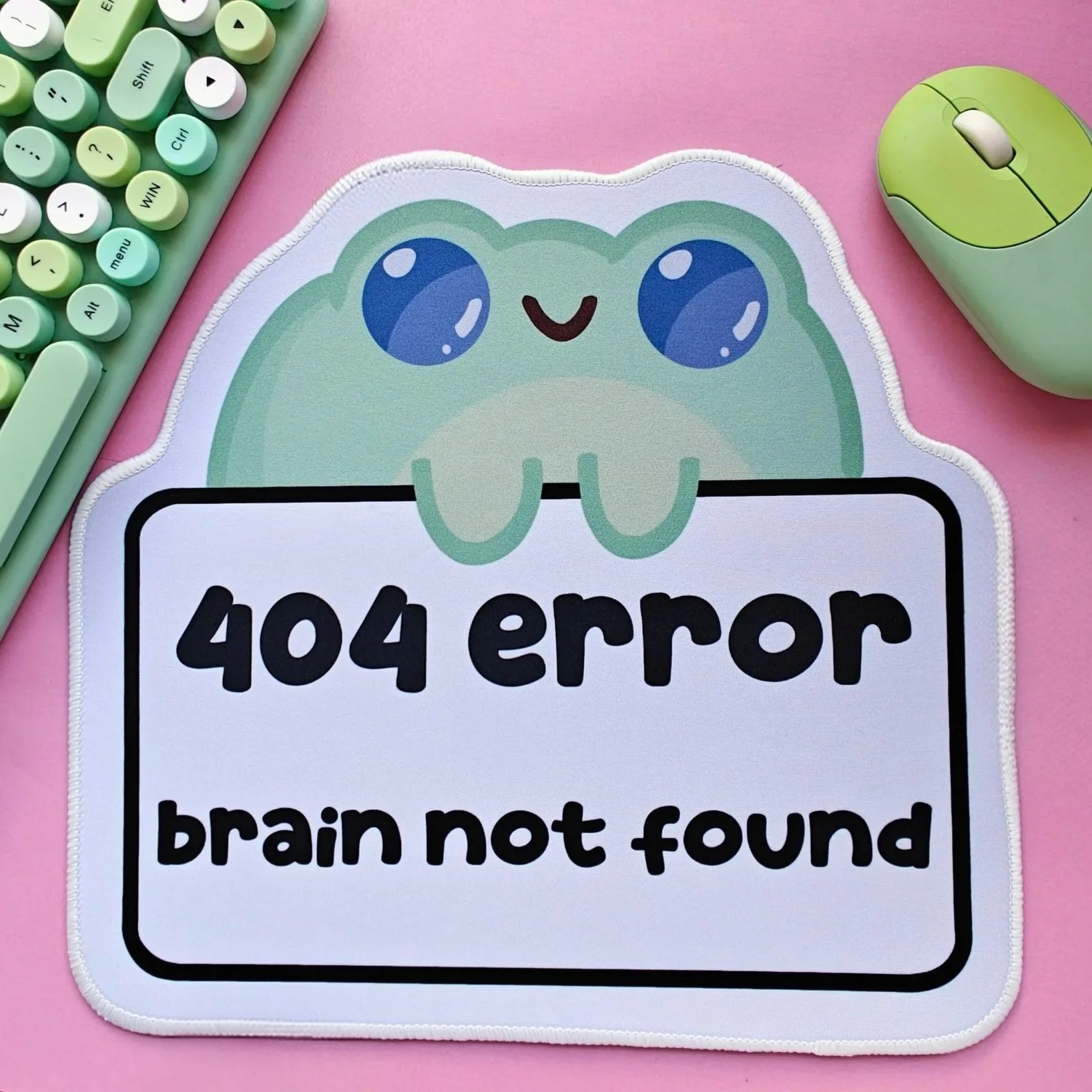 A light green cartoon frog mousemat with large blue eyes and a happy face, holding a sign that reads '404 error brain not found' on a white background. The scene includes a mint green keyboard and mouse on a pink surface.