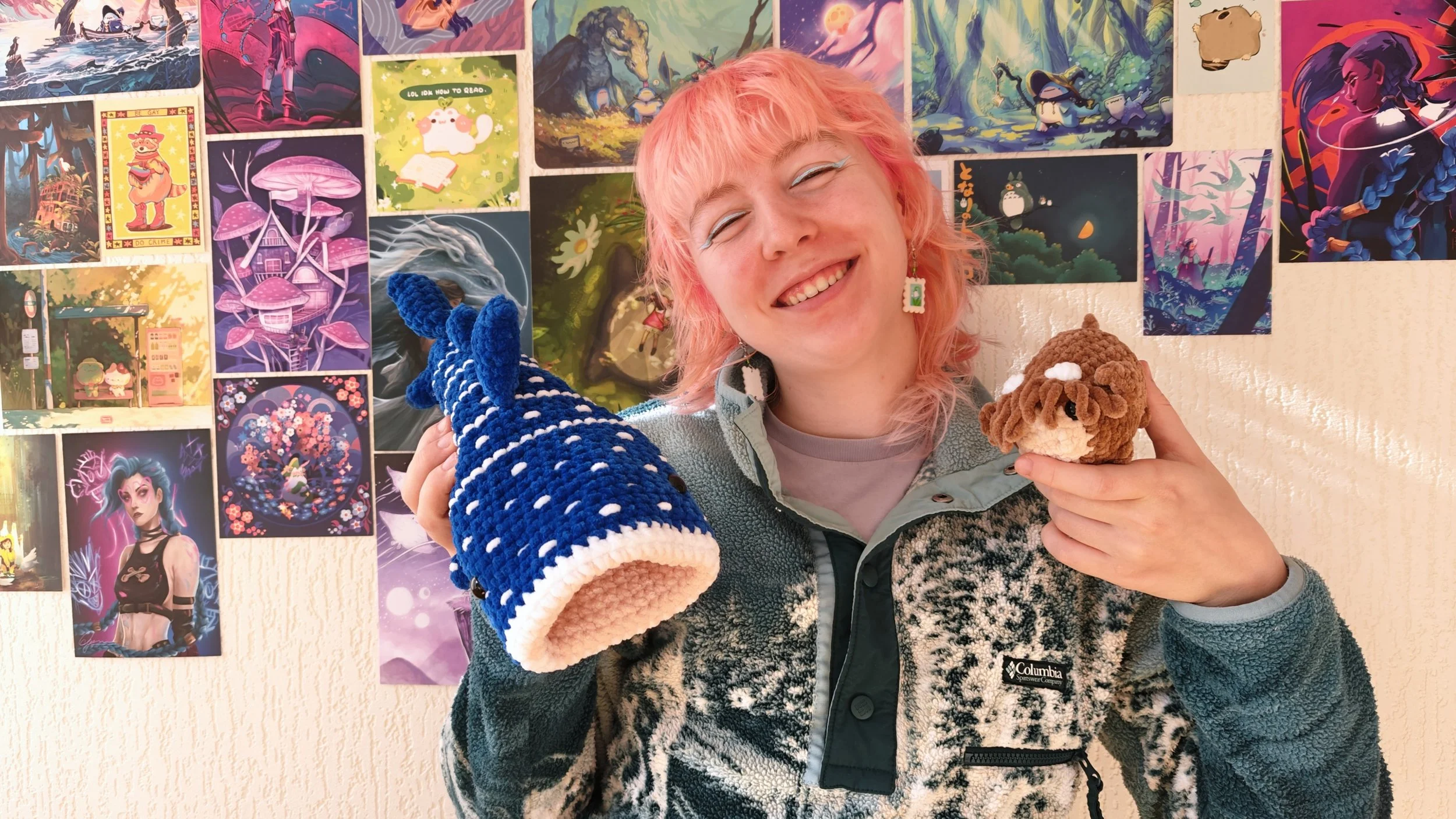 A person with pink hair smiling while holding a blue crocheted whale and a small plush cow in front of a colorful wall of various illustrations.