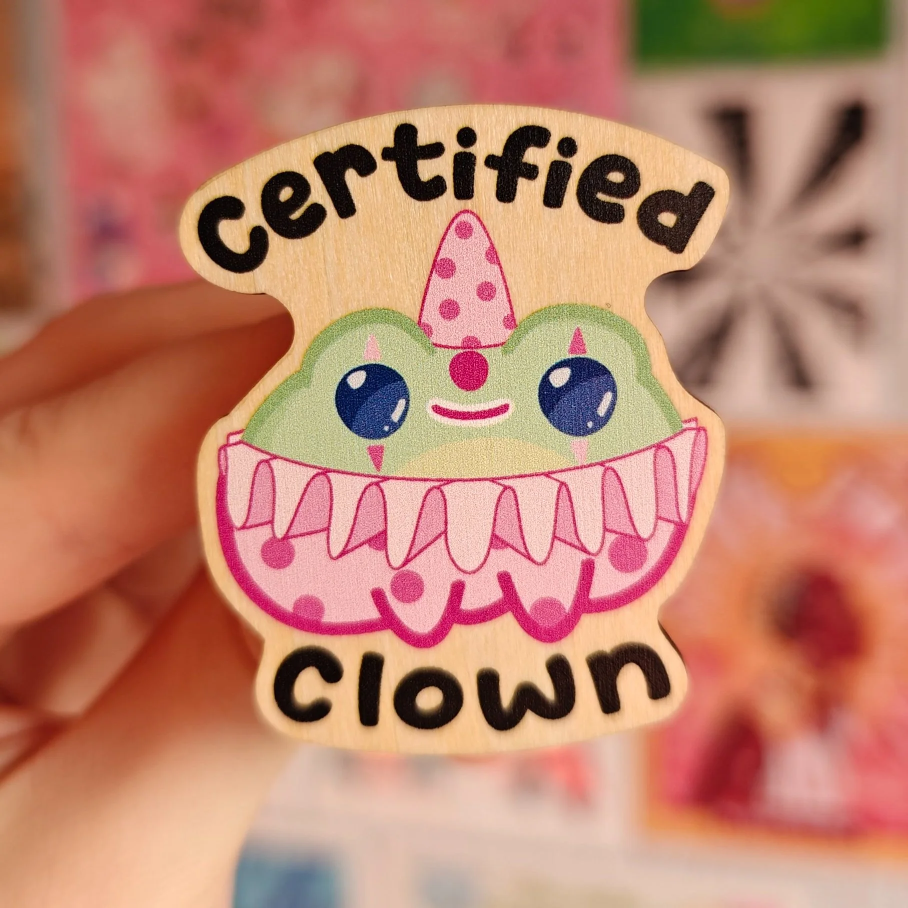 A sticker with the words 'certified clown' and a cartoon frog with big eyes, a pink party hat, and a pink ruffled collar.