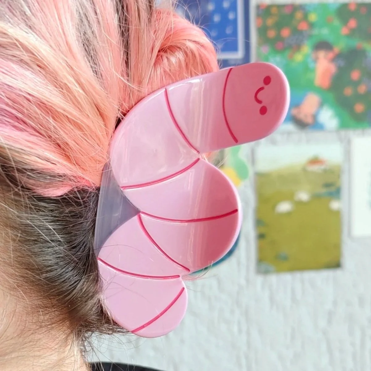Pink worm hair claw with a smiling face design in a person's hair, with colorful children's drawings in the background.