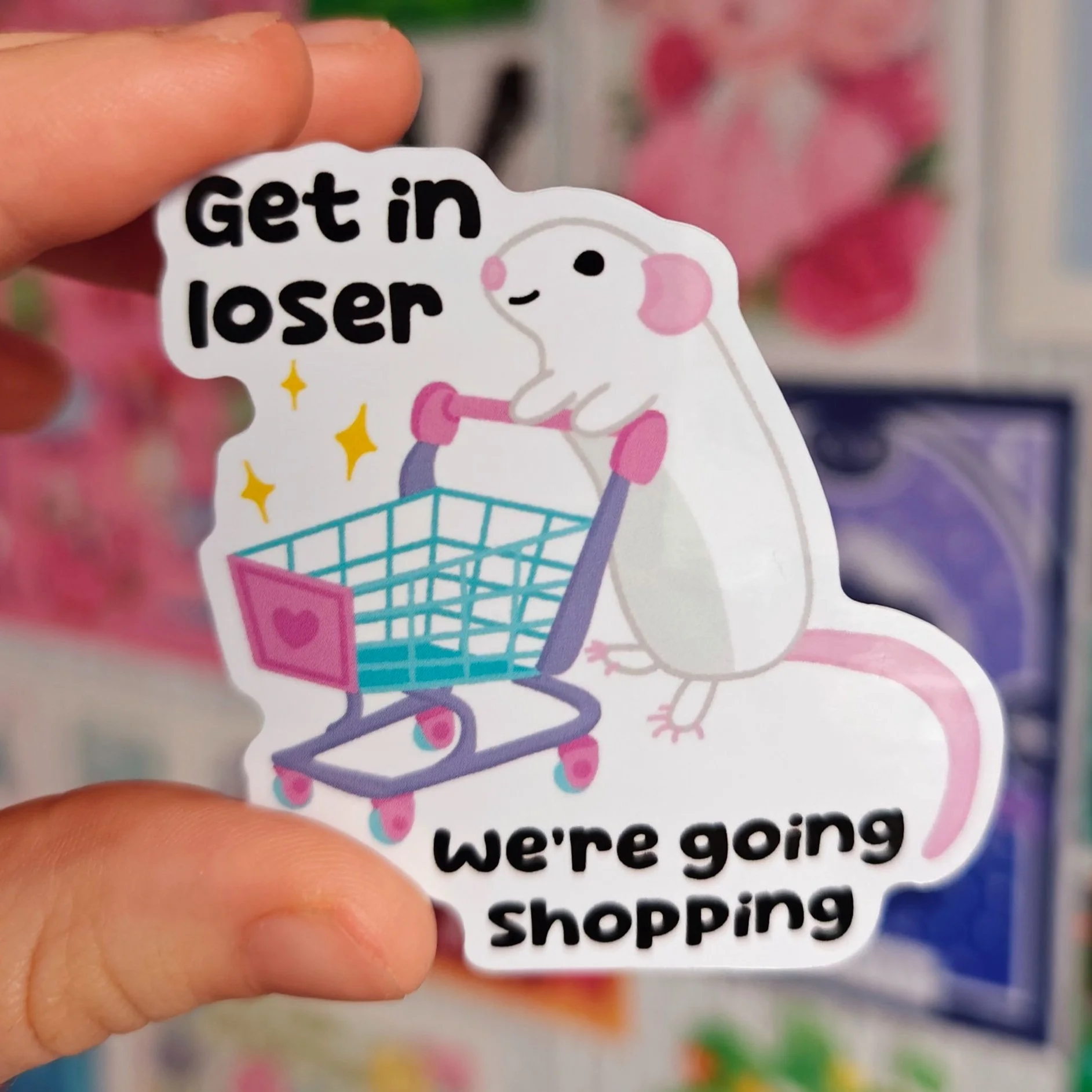 A sticker featuring a cute cartoon white mouse with pink ears and tail, pushing a shopping cart with a pink heart and yellow sparkles, accompanied by the playful text 'get in loser we're going shopping'.