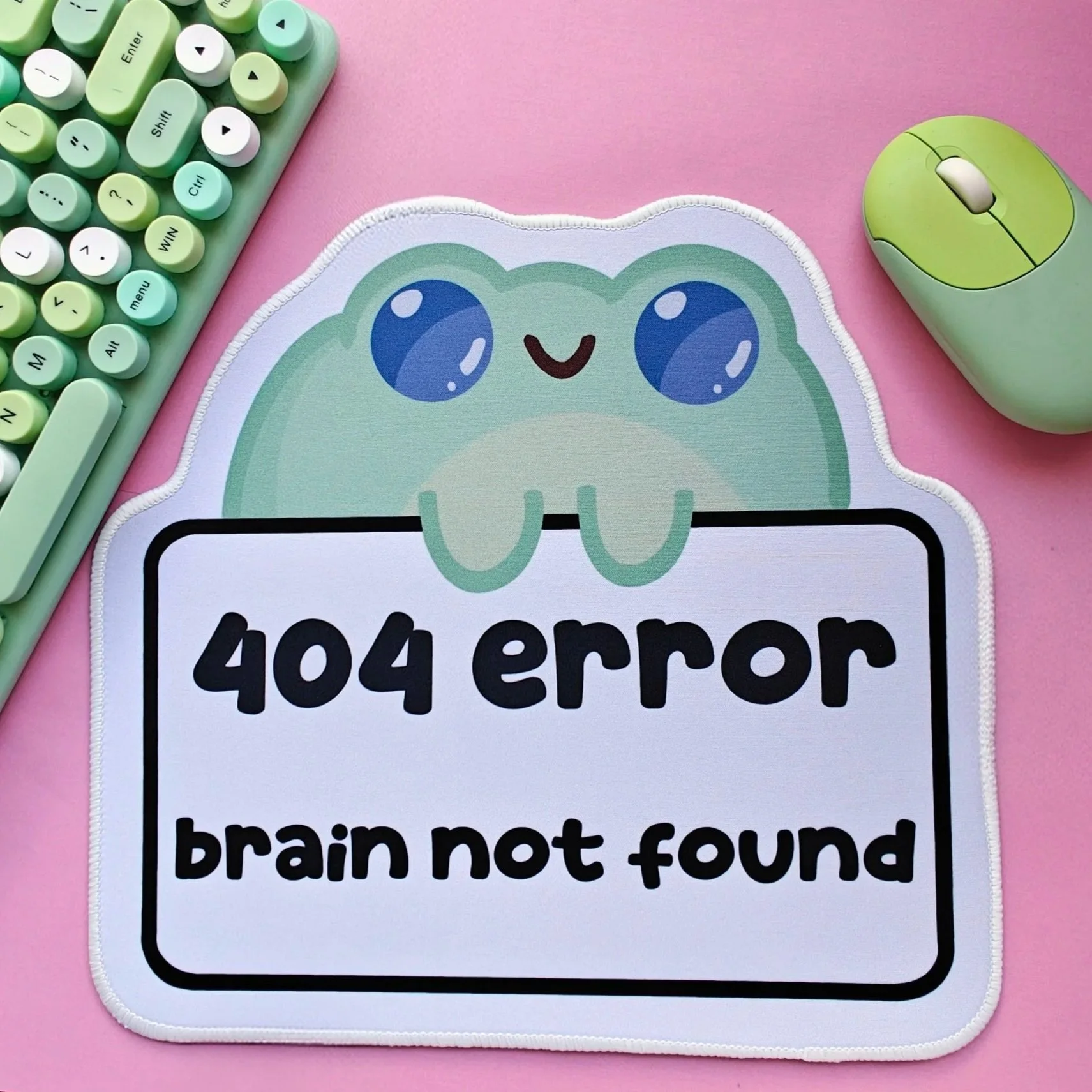 A mouse pad with a cartoon frog and the text '404 error brain not found', placed on a pink surface. To the left, a mint green keyboard with round keys, and to the right, a matching mint green computer mouse.