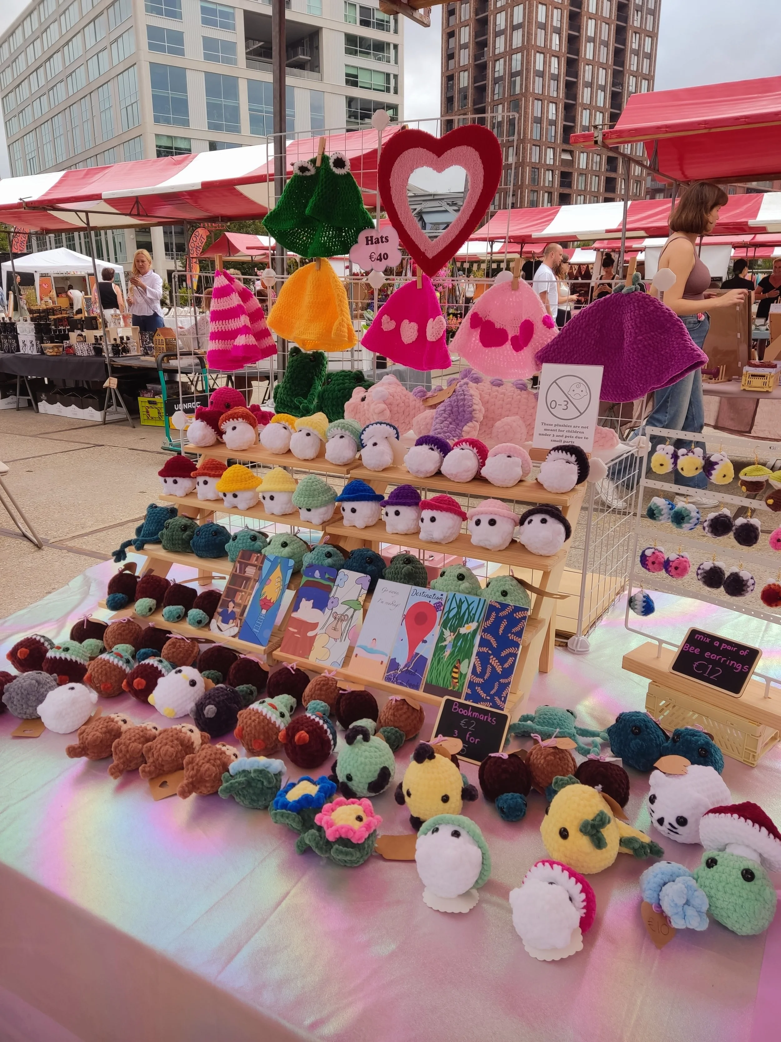 A market stall displays colorful handmade hats, plush toys, and bookmarks, with city buildings in the background.