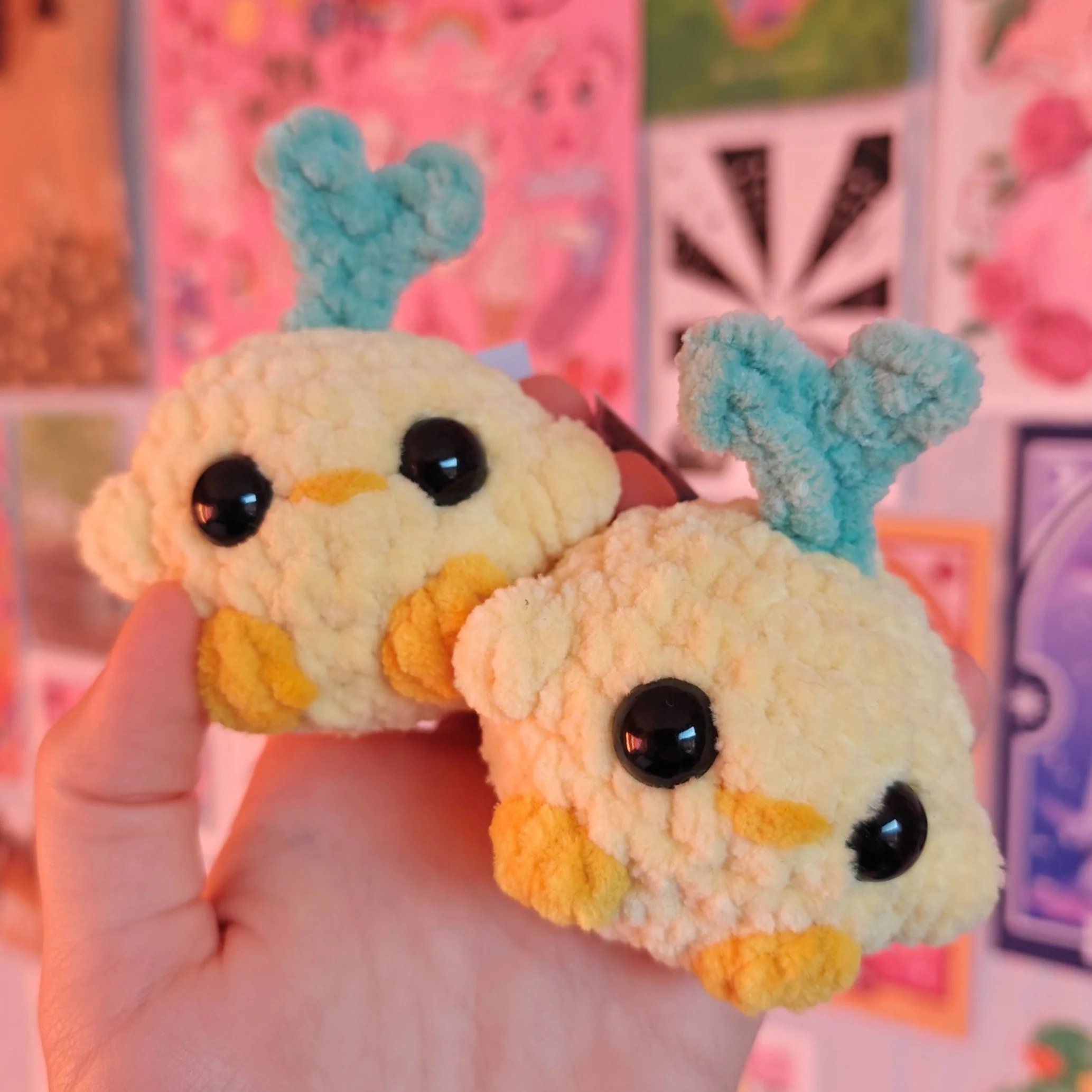 Two plush duck toys with large black eyes, orange beaks, and blue yarn-combed necks, held in a hand against a colorful background.