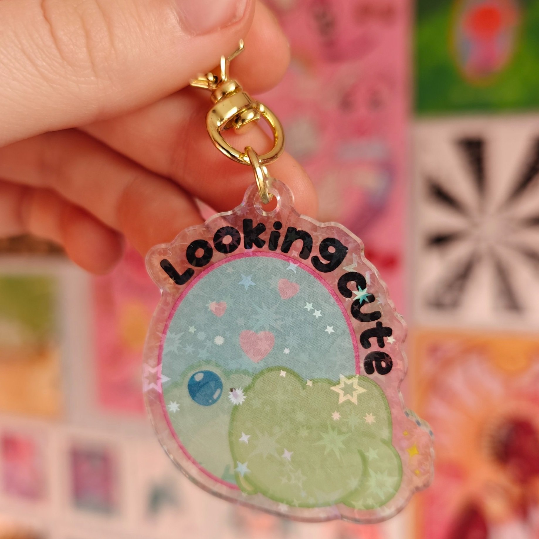 Close-up of a hand holding a colorful keychain with the words 'Looking Cute' and a cartoon frog design in the background.