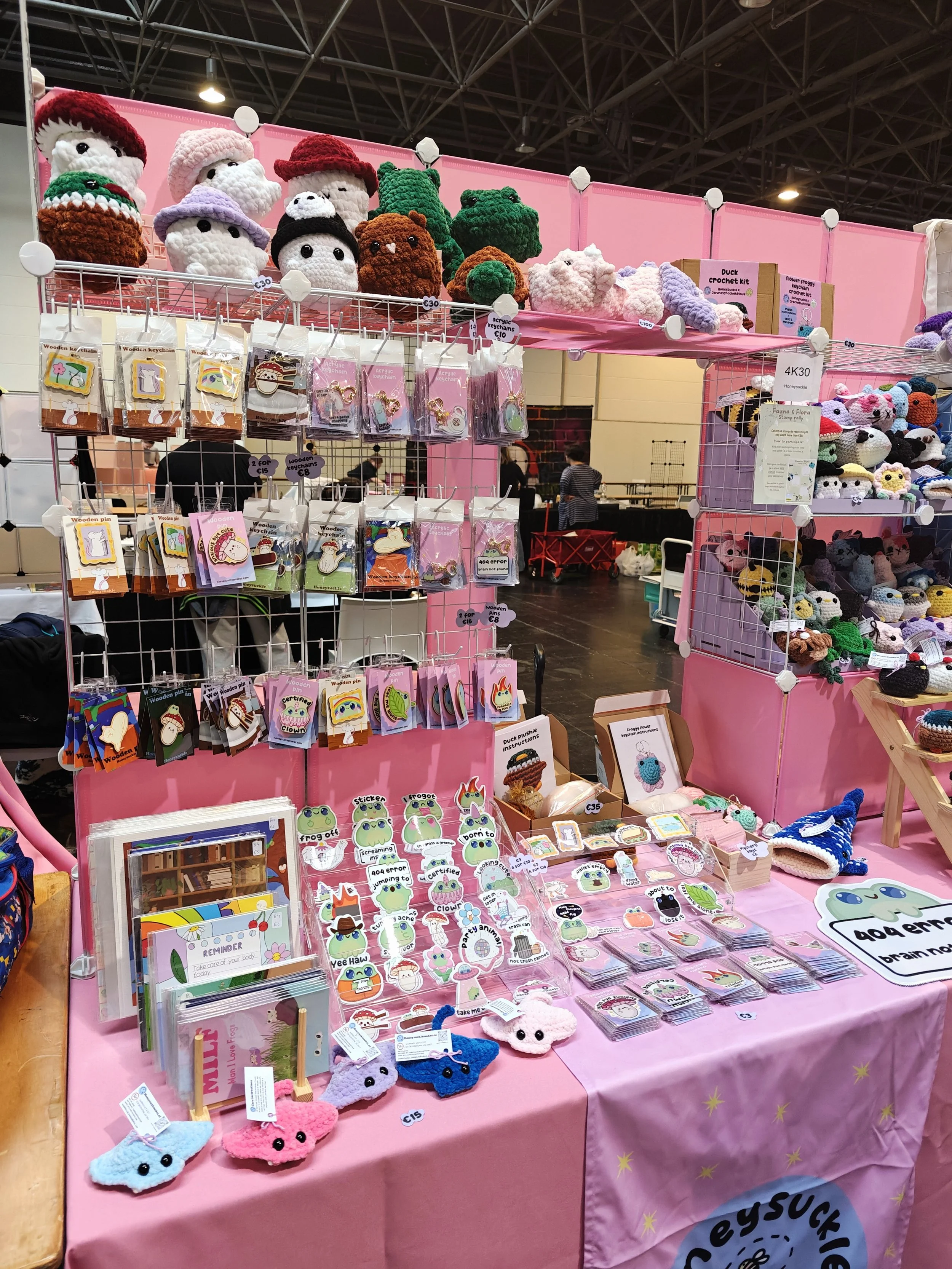 A display booth with various handmade crochet and craft items, including stuffed animals, keychains, and bookmarks, set up on pink tables and shelves at a comic convention.