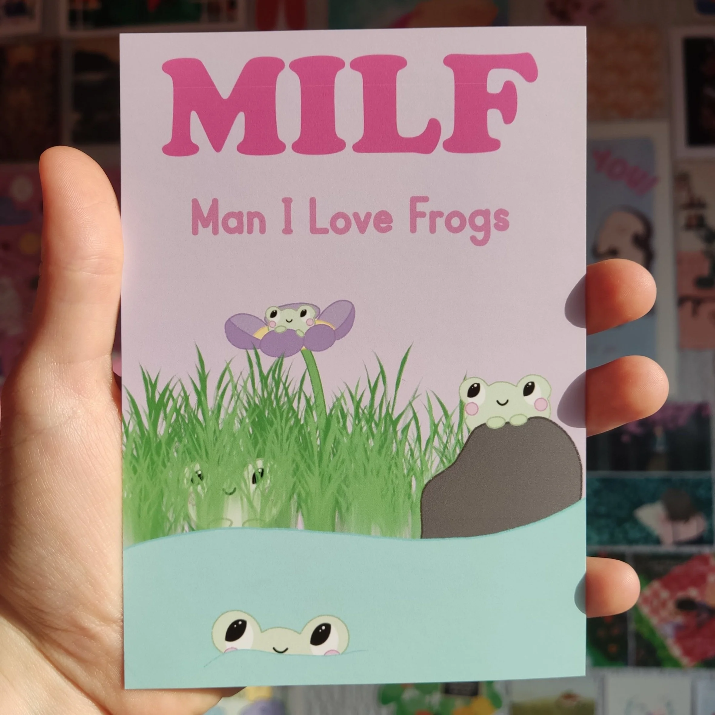 A pink and light purple card with the word 'MILF' at the top, and the phrase 'Man I Love Frogs' underneath. The illustration features cute cartoon frogs among grass, rocks, and a purple flower, with some frogs peeking out from water.