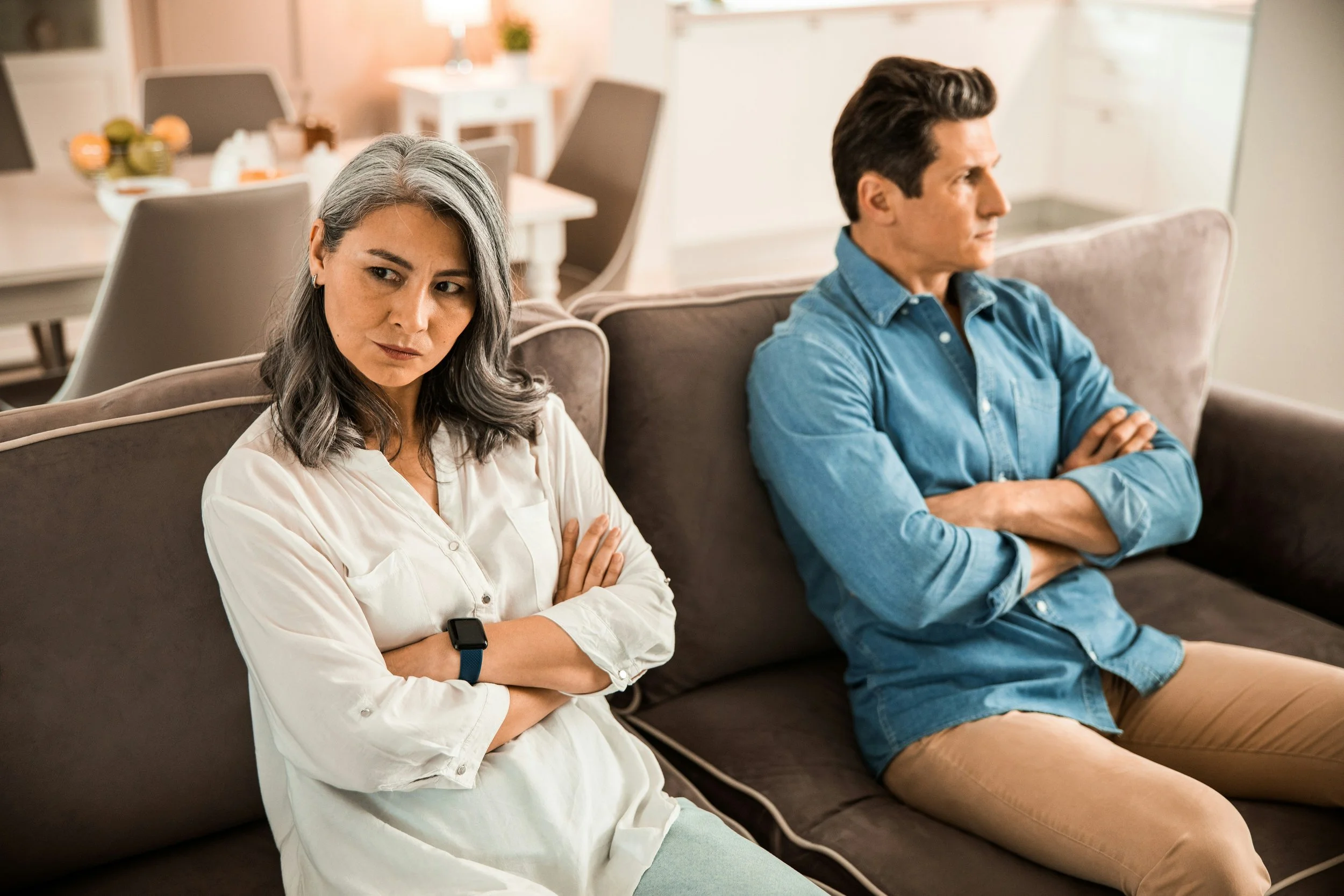 Partners sitting apart with crossed arms during conflict, representing how unresolved trauma affects relationships and how couples counseling trauma work can support repair and reconnection.